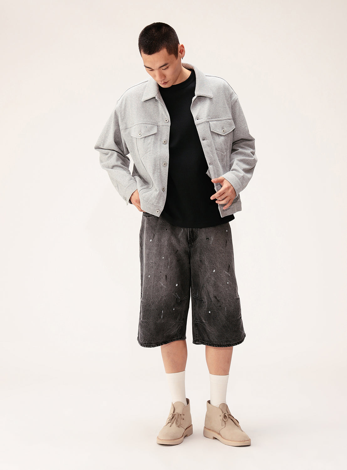 A person stands against a plain light background wearing the BEAMS Button-Down Jersey Trucker Jacket over a black shirt, dark gray splatter-pattern shorts, white socks, and beige desert boots.
