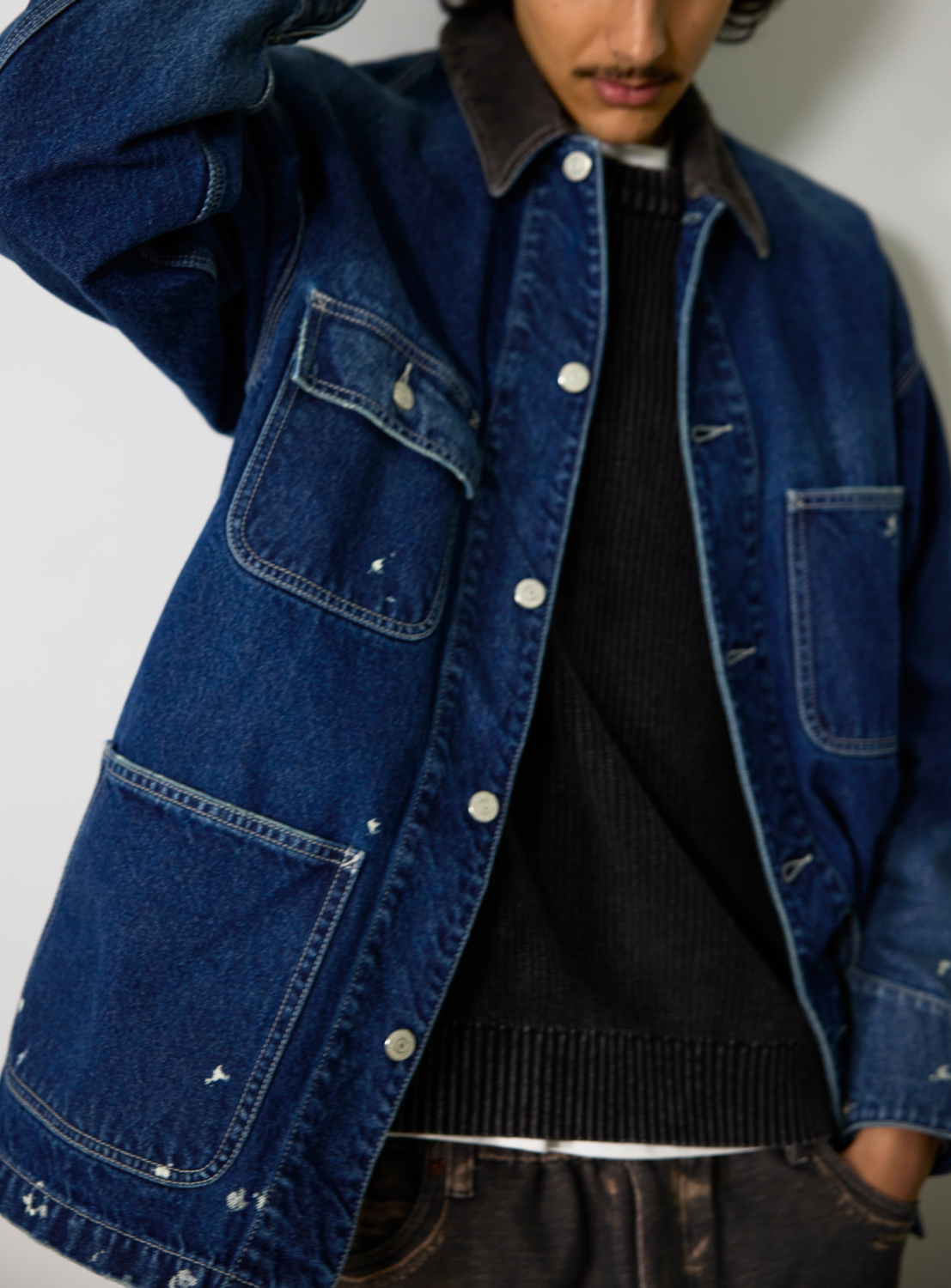 A person wearing a BEAMS Used Denim Coverall featuring a vintage, splash-bleached wash, dark corduroy collar, and buttoned chest pockets over a black sweater.