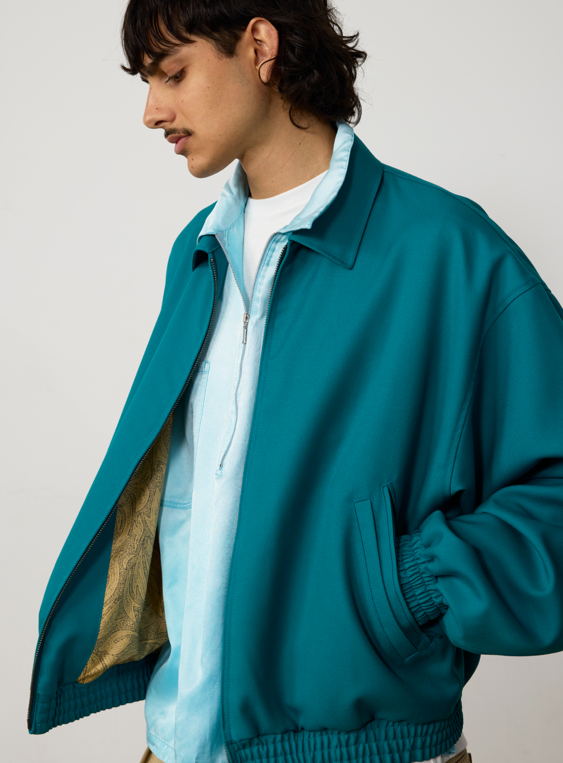 A person wearing an unzipped teal wool BEAMS Gabardine Zip Blouson over a light blue collared shirt.