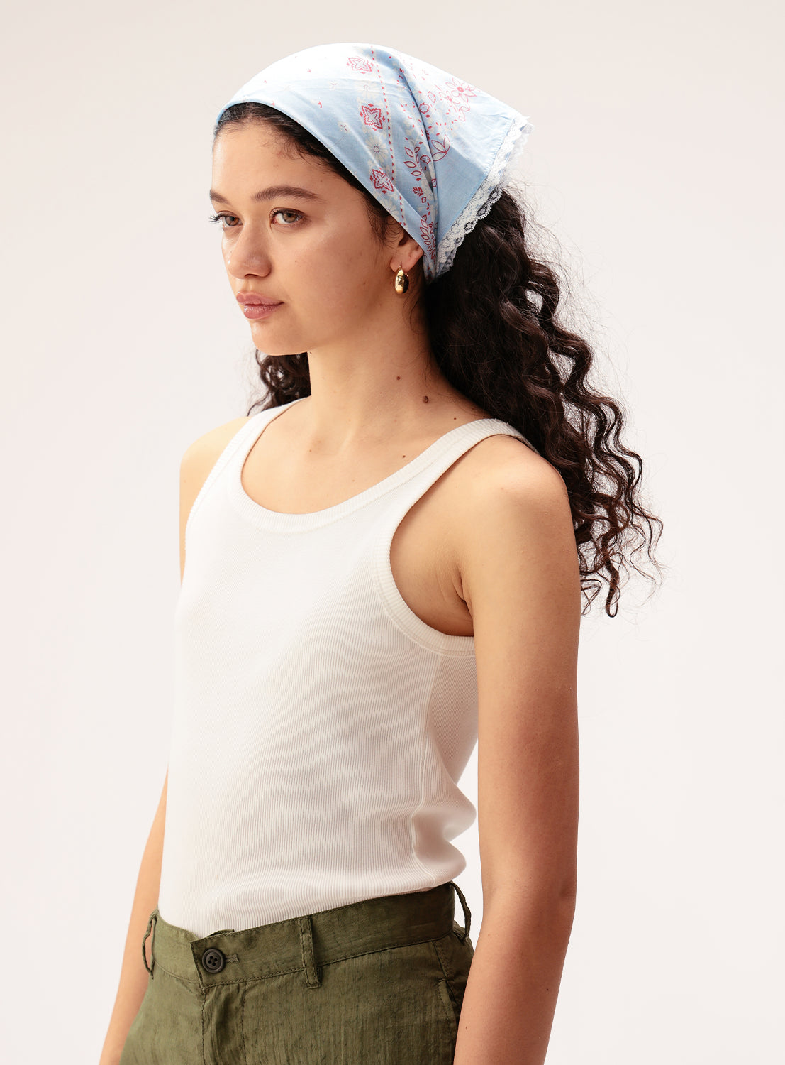 A young woman with long curly hair wears the BEAMS BOY 2-Way Floral Lace Bandana, styled vintage-inspired in light blue with delicate floral patterns, paired with a white tank top and green pants as she stands against a plain background.