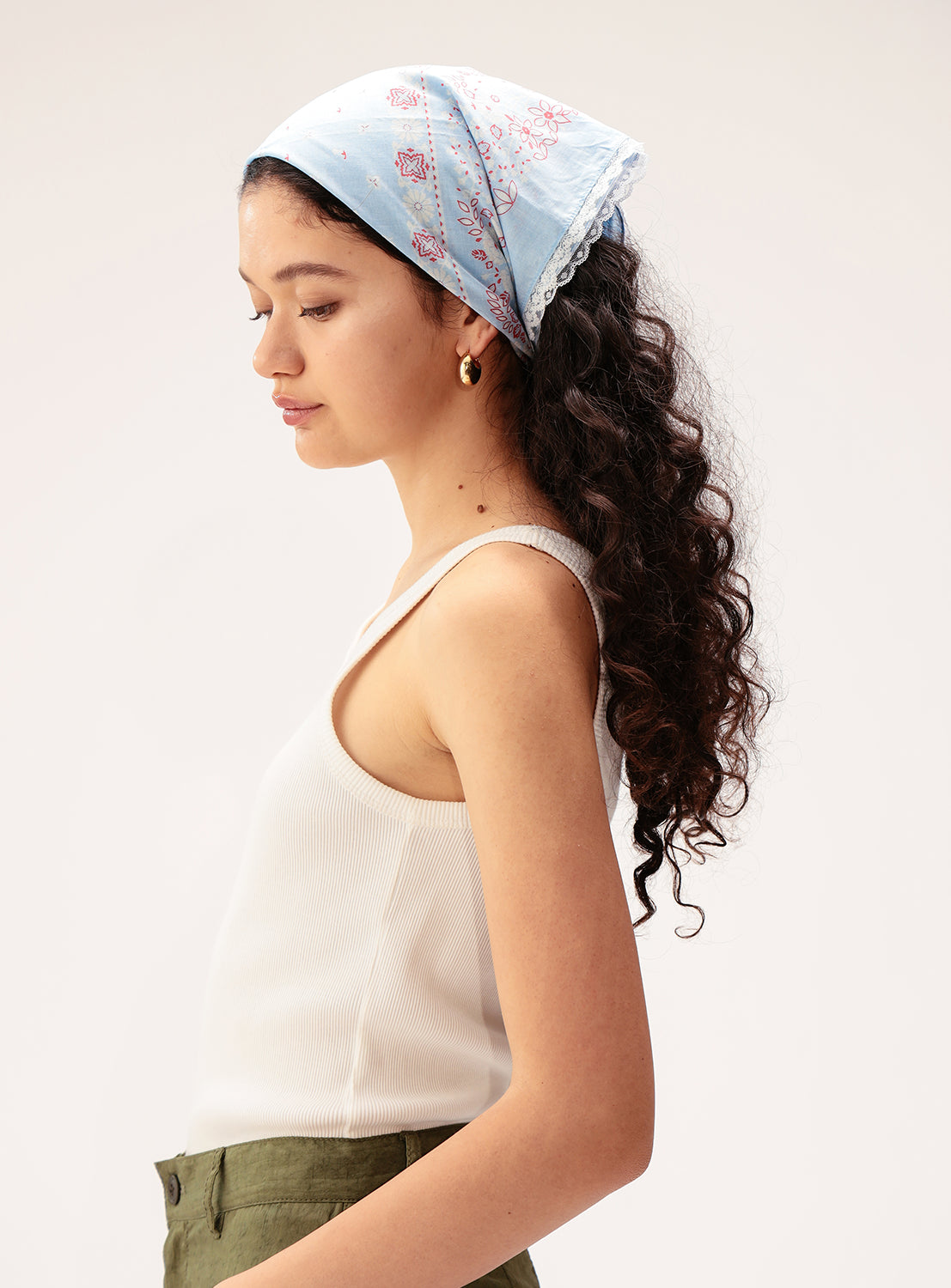 A woman in profile wears a white tank top, olive green pants, and the BEAMS BOY 2-Way Floral Lace Bandana—a vintage-inspired blue patterned accessory with delicate trim. She has long, curly hair and gold hoop earrings, standing against a plain background.