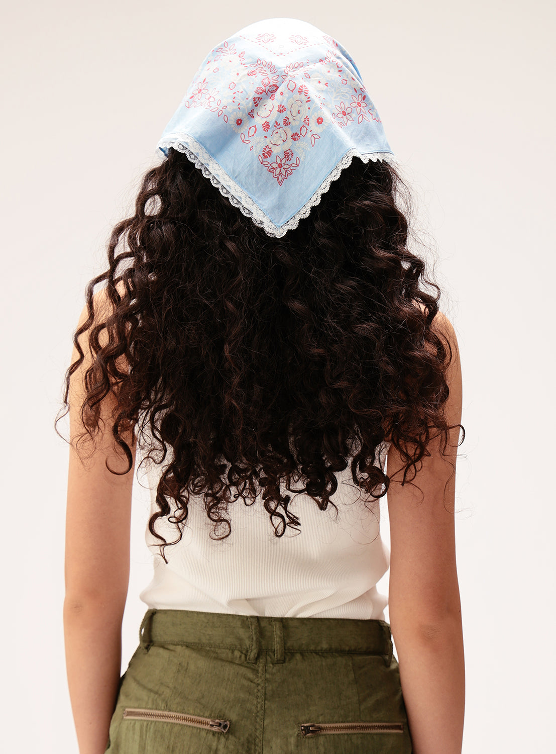 A person with long, curly dark hair is shown from the back wearing a white tank top, olive green pants, and the BEAMS BOY 2-Way Floral Lace Bandana—a vintage-inspired light blue scarf with a pink pattern and lace trim.