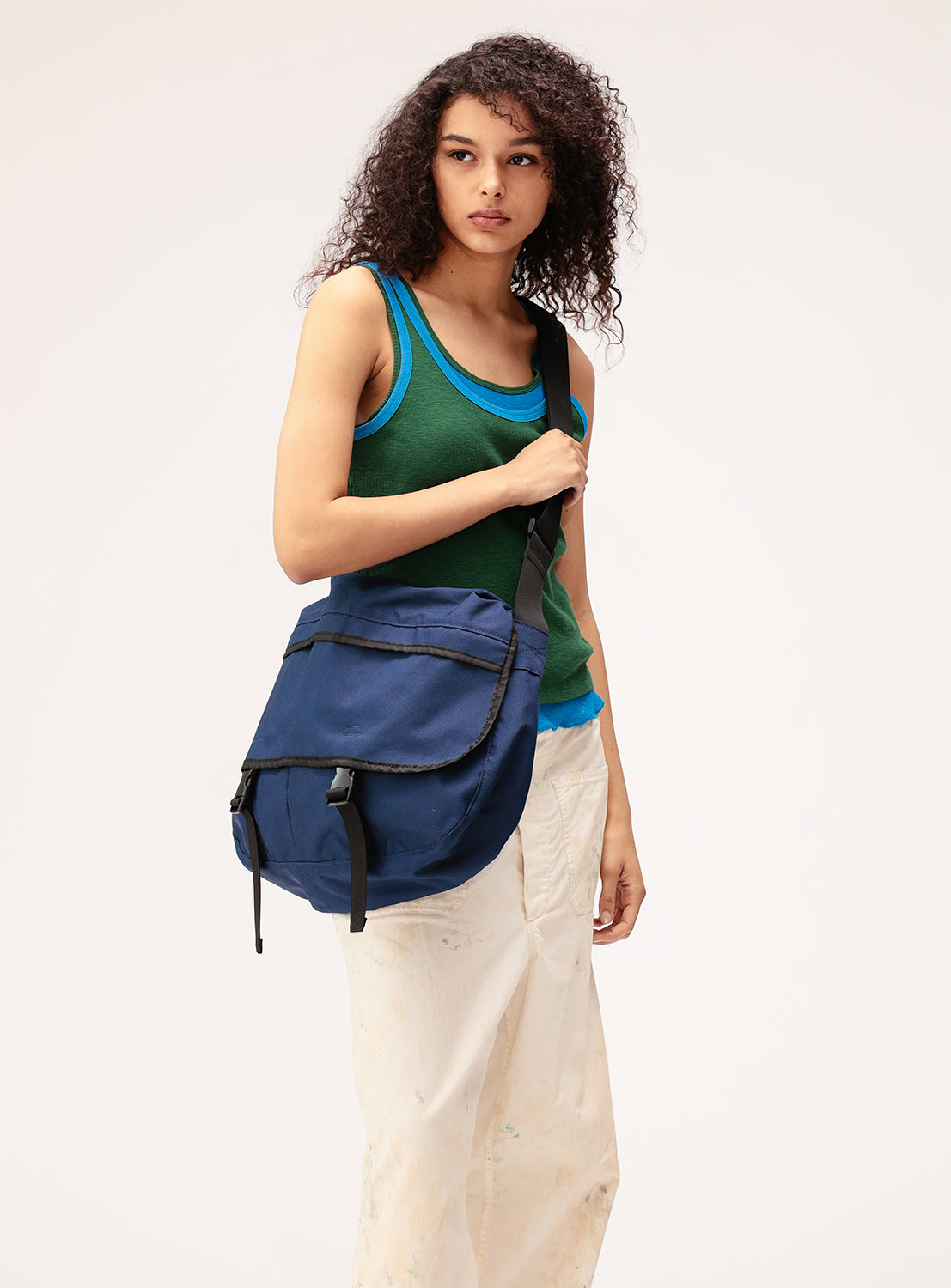 Wearing a green tank top and white pants, a person with curly hair stands against a plain light background, carrying the BEAMS BOY Oversized Messenger Bag for a statement silhouette.