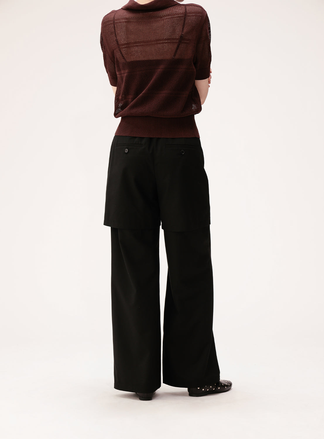 A person stands facing away against a plain light background, wearing a sheer maroon short-sleeve top, black shorts layered over Ray BEAMS Half Layered Wide-Leg Trousers crafted from lightweight suiting fabric, and black shoes.