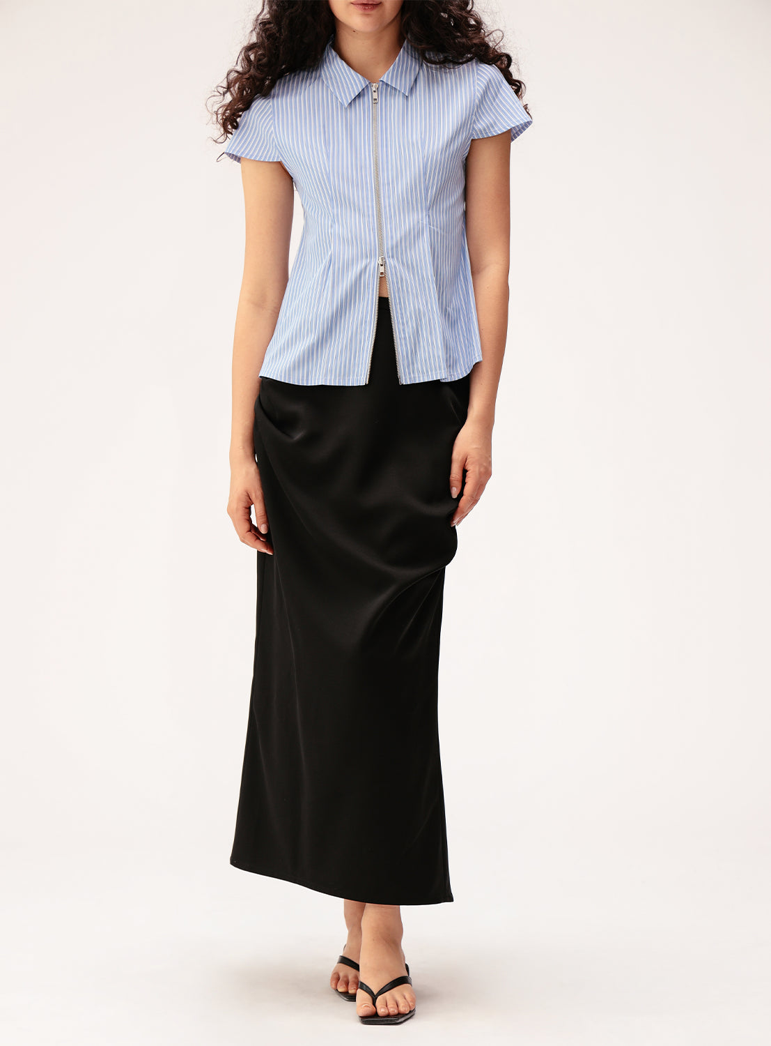 A woman wearing a short-sleeved, blue striped button-up top and the Ray BEAMS Satin Drape Skirt with sculptural front detail, paired with black sandals, stands against a plain white background.