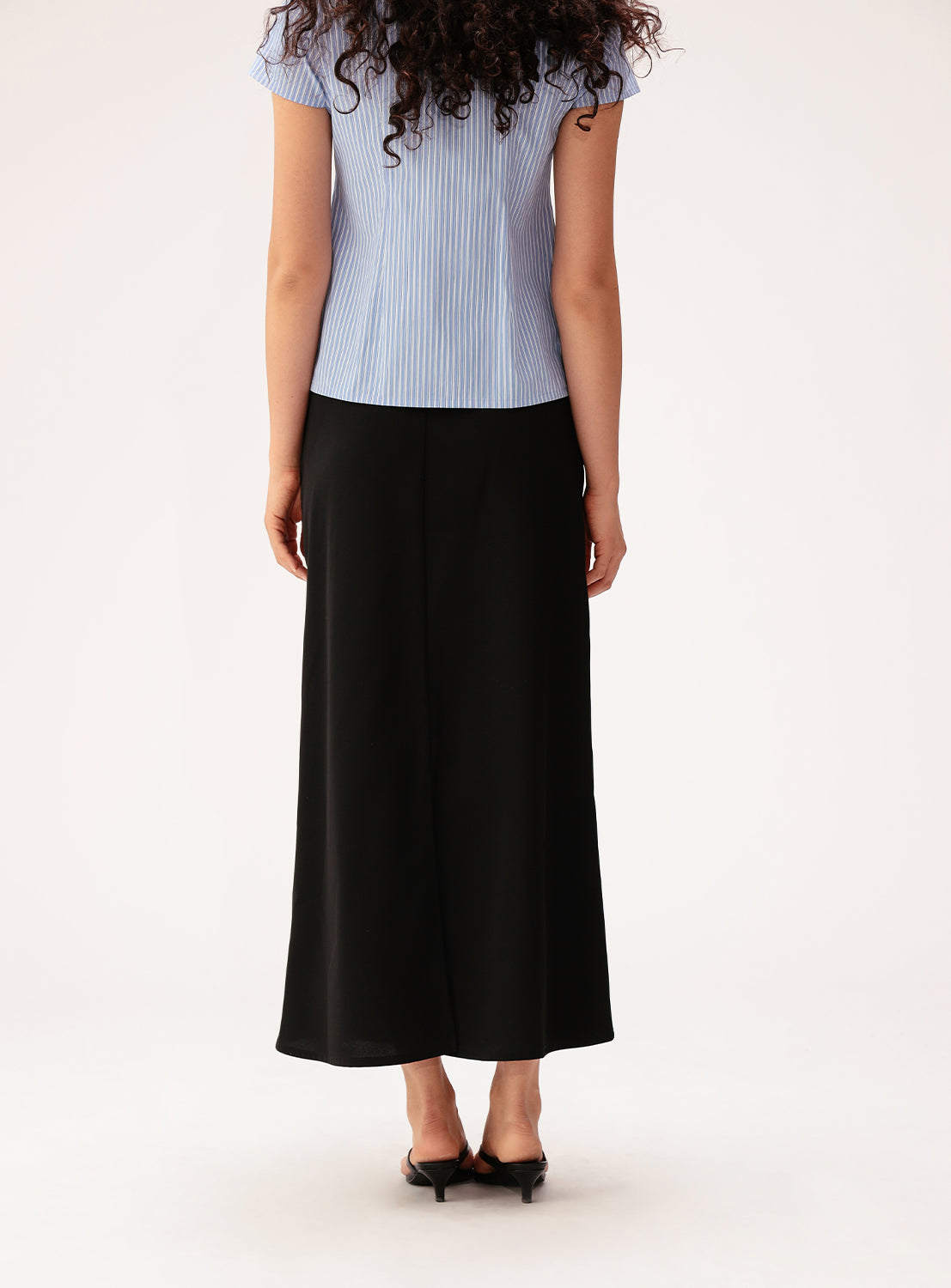 A woman with long curly hair stands facing away, wearing a short-sleeved blue striped blouse paired with the Ray BEAMS Satin Drape Skirt—an elegant black skirt featuring a sculptural front detail—and black heels against a plain, light background.