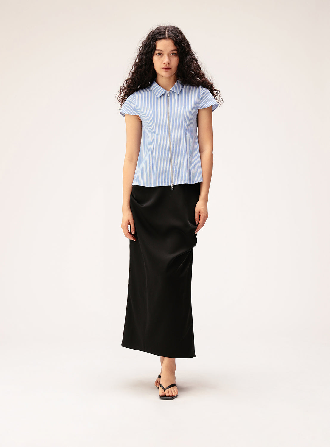 A woman with long curly hair wears a blue short-sleeve button-up shirt and the Ray BEAMS Satin Drape Skirt in black with sandals, standing against a plain light background.