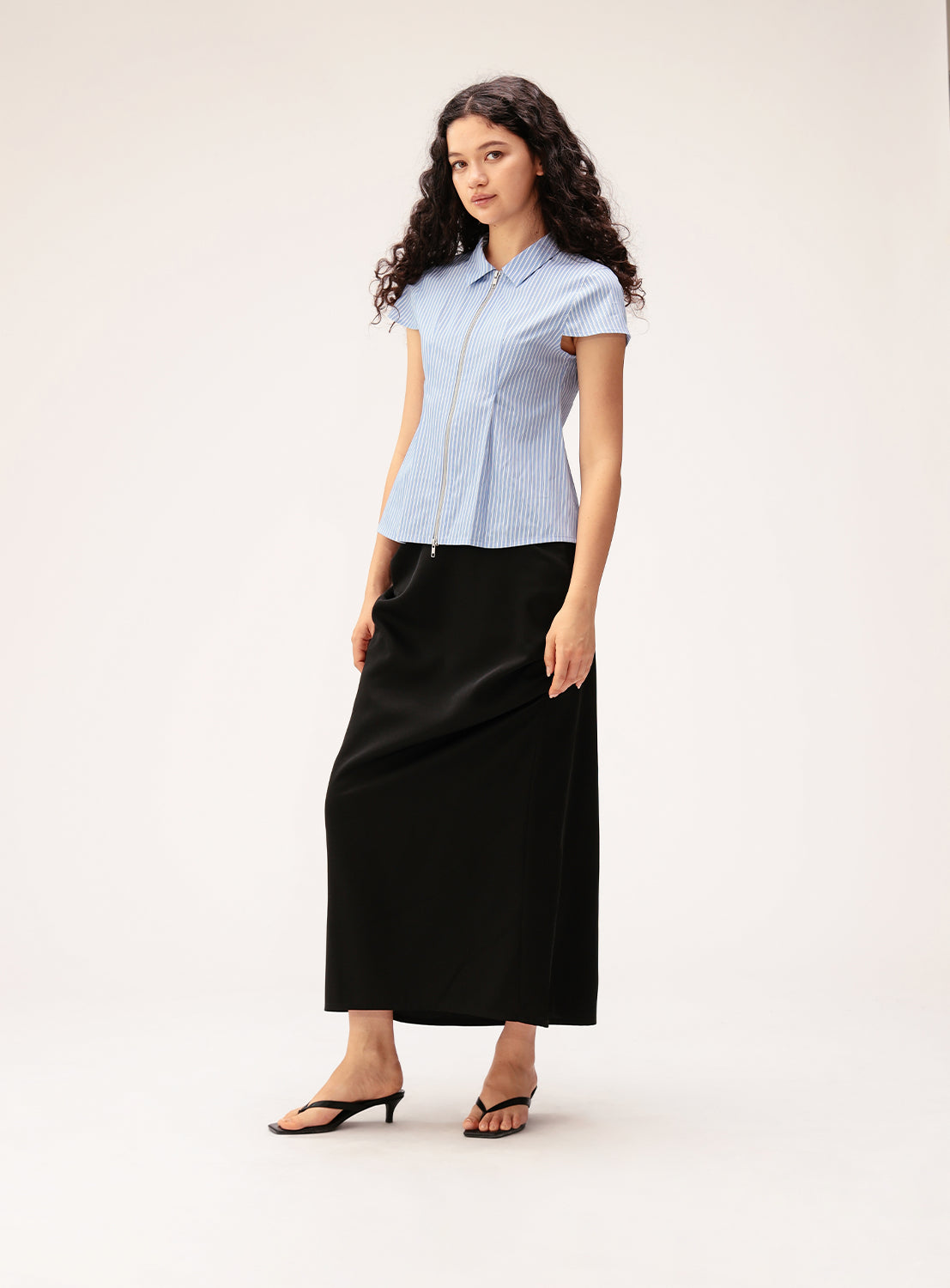 A woman with long, curly hair wears a short-sleeve blue button-up shirt, the Ray BEAMS Satin Drape Skirt—an elegant black ankle-length skirt featuring sculptural front detail—and black sandals, standing against a plain light background.
