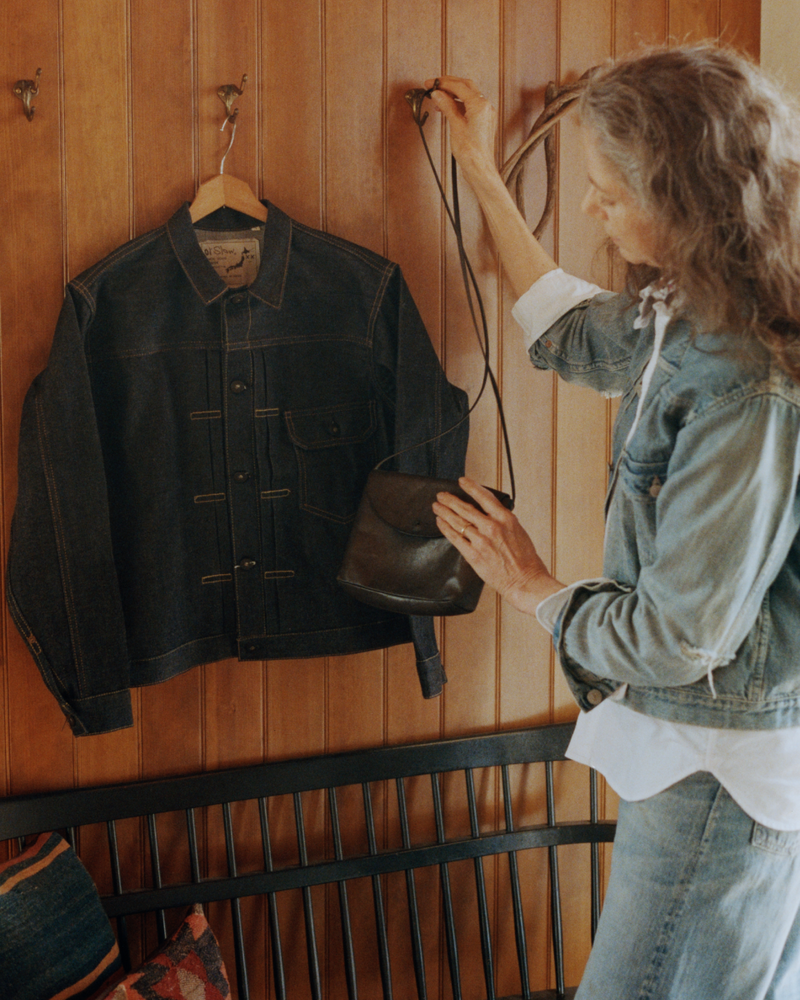 An older woman in a denim jacket and jeans hangs a small black purse on a hook beside a hanging dark denim jacket, against a wooden wall.