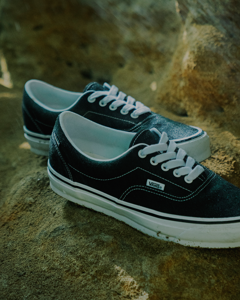 A pair of black Vans sneakers with white laces and soles sit on a textured, rocky surface in natural light.