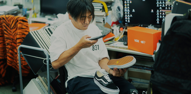 A man in a white shirt sits at a cluttered desk, examining a sneaker and taking a photo of it with his phone. Computer monitors, shoe boxes, and stacks of papers fill the busy office space around him.