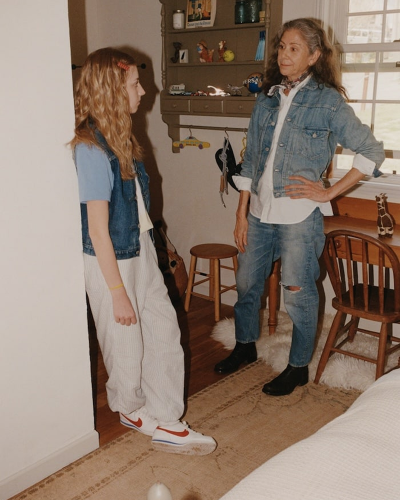 An older woman and a girl stand talking in a cozy room. Both wear denim jackets. The woman stands near a wooden chair, and the girl leans against the wall. The room has warm lighting and homey decorations.