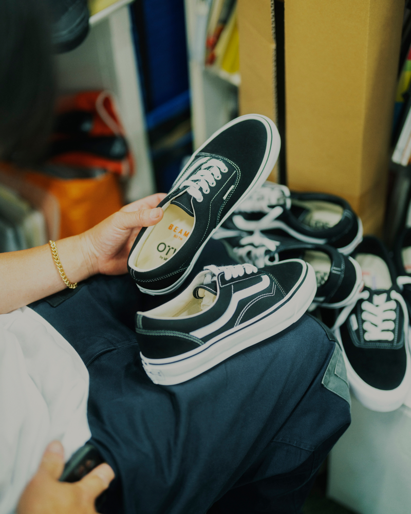 A person holding a black sneaker with white laces, sitting with several similar sneakers on their lap and nearby. Shelves, boxes, and books are visible in the background.