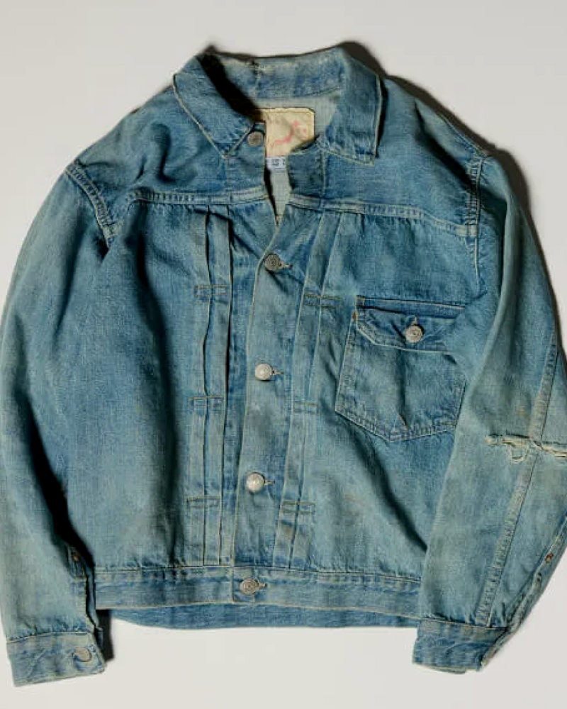 A faded blue denim jacket with metal buttons, a collar, and a single chest pocket is laid flat on a white background.