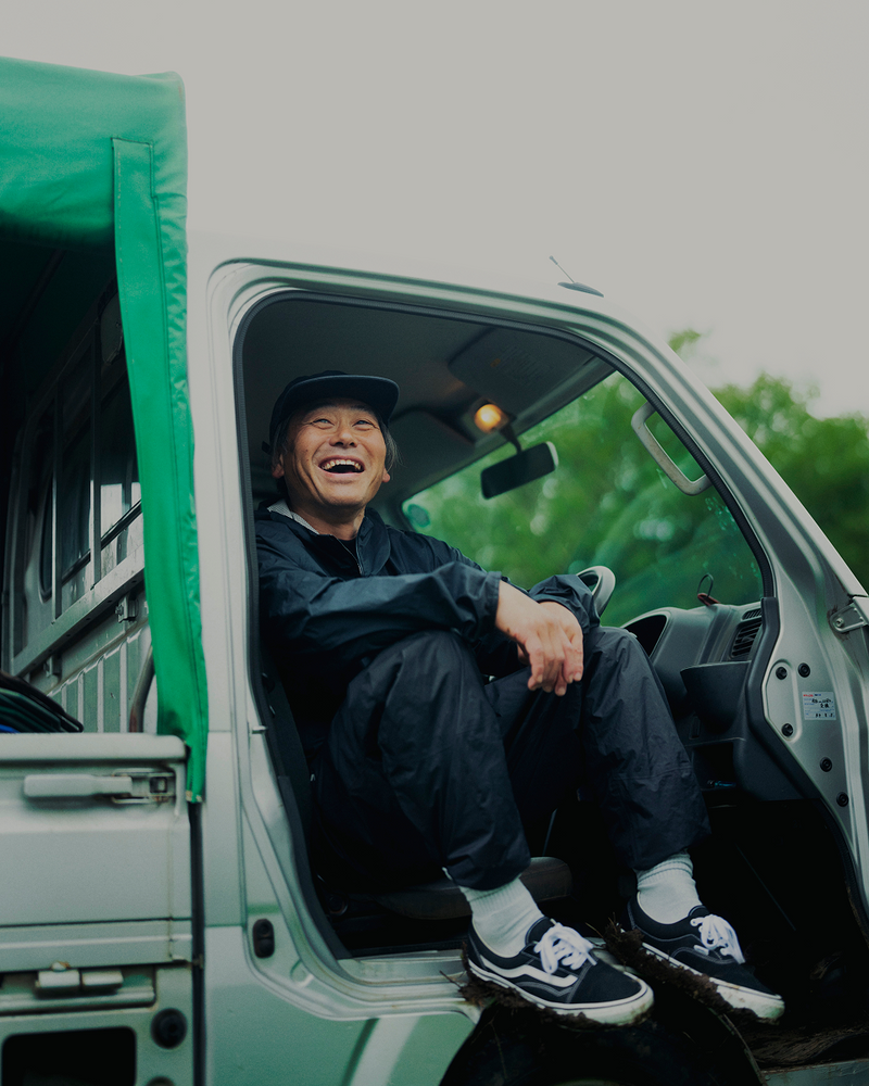 A smiling person wearing a cap and dark jacket sits in the open doorway of a vehicle with a green cover, feet on the step, and greenery visible outside.