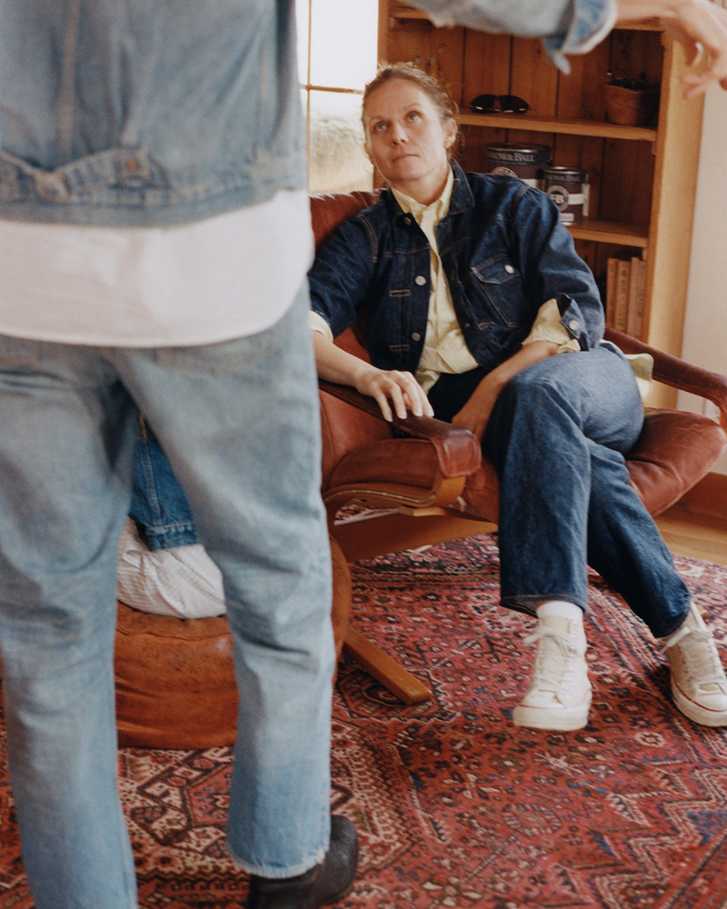 A person in a denim jacket and jeans sits in a chair, looking up at another person standing nearby. The setting is indoors with a patterned rug and shelves in the background.