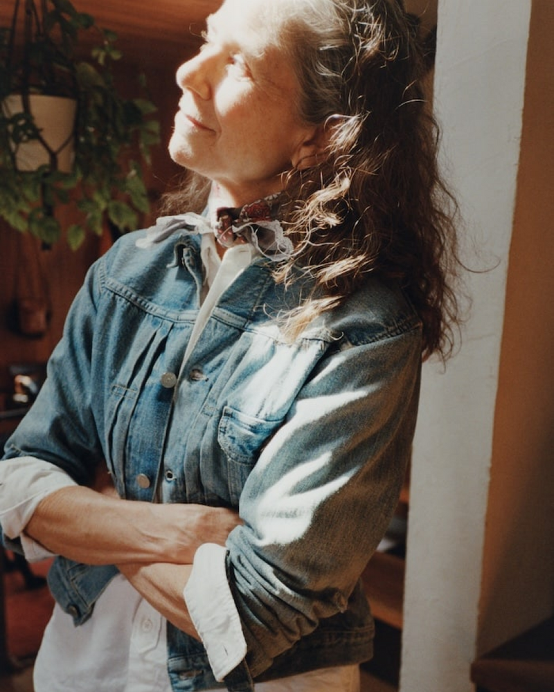 A woman with long wavy hair stands with arms crossed, wearing a denim jacket and scarf, looking up into sunlight streaming through a window in a warmly lit room.