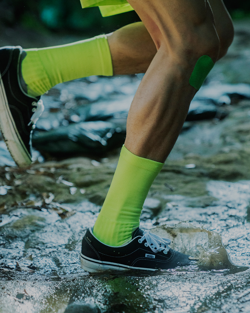 A person wearing neon yellow socks, black sneakers, and green kinesiology tape on their calf steps onto a wet, muddy surface outdoors.