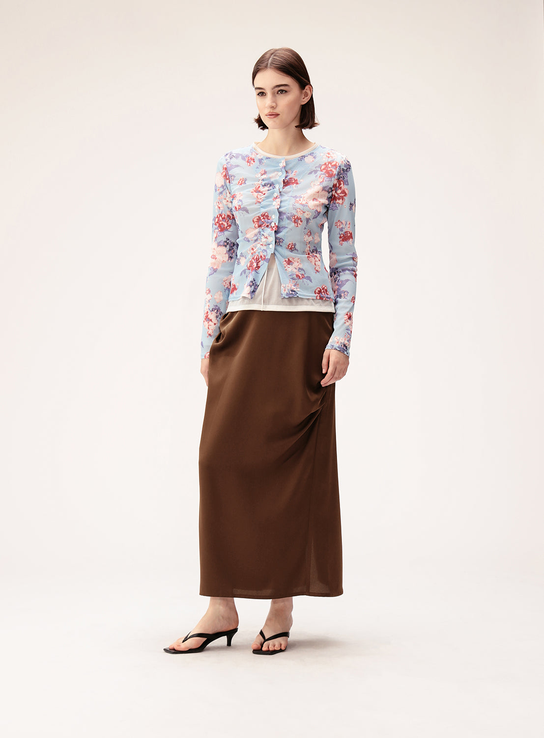 A woman stands against a plain, light background wearing a light blue floral cardigan over a white top, paired with the Ray BEAMS Satin Drape Long Skirt in brown and black sandals.