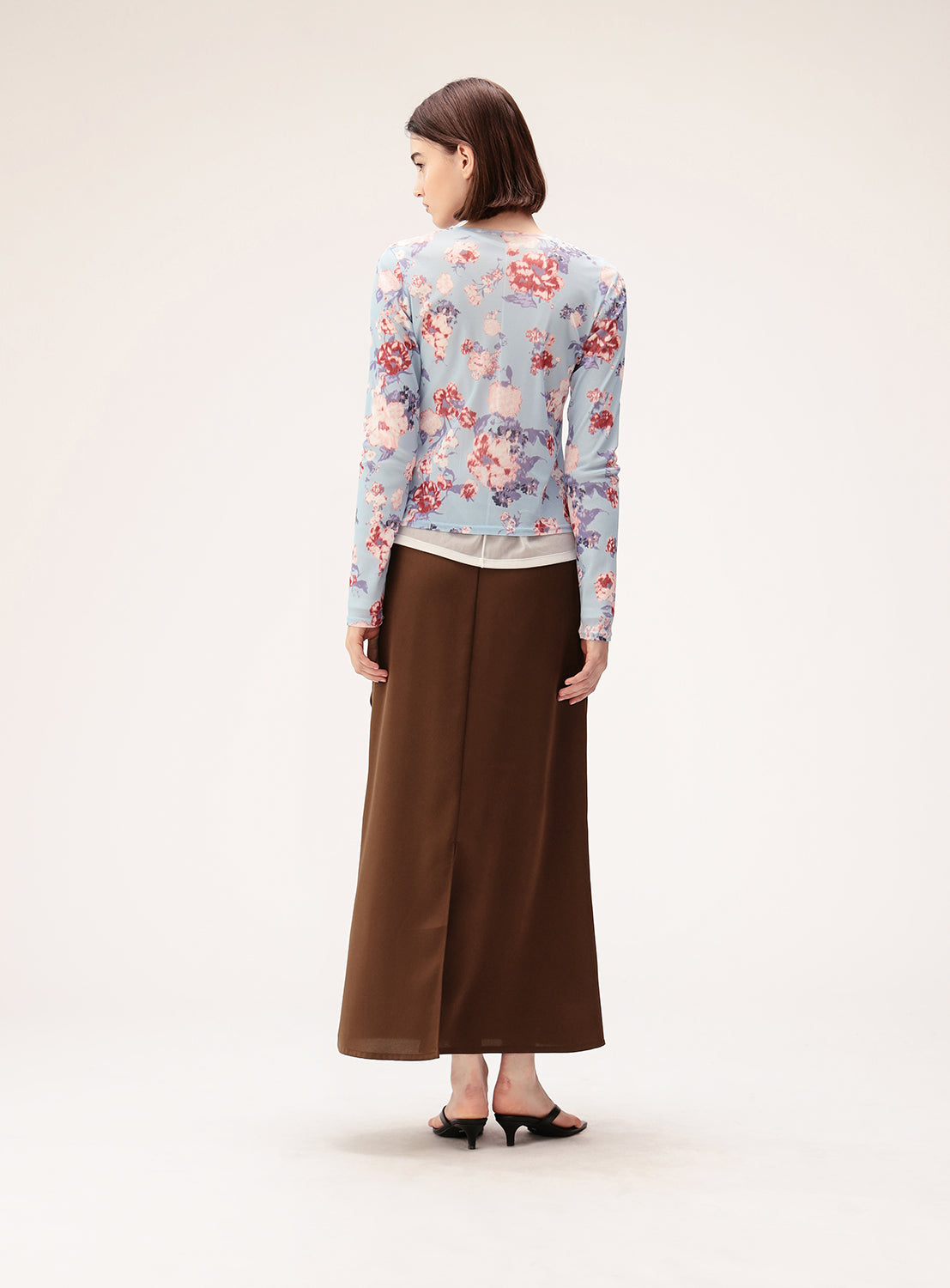 A woman stands facing away, wearing a light blue floral long-sleeve top paired with the Satin Drape Long Skirt by Ray BEAMS and black heeled sandals, set against a plain light background.