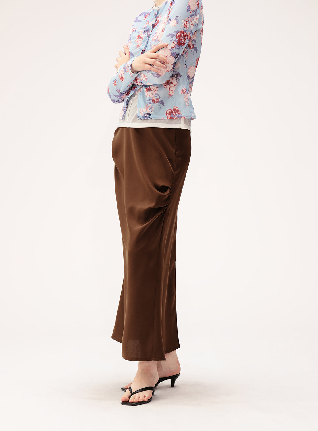 A person wearing a light blue floral top, the Ray BEAMS Satin Drape Long Skirt with a versatile silhouette, and black heeled sandals stands against a plain white background with arms crossed.