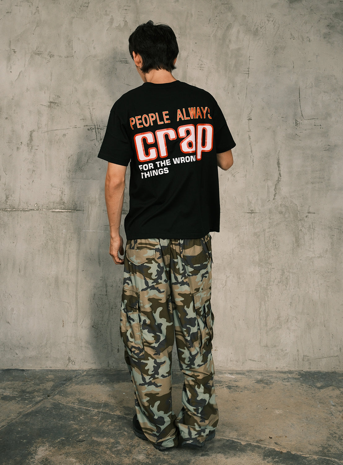 Facing a textured gray wall, the individual wears the BIAS DOGS 'CRAP' Graphic Tee—featuring the bold statement "People Always Crap For The Wrong Things"—paired with camouflage cargo pants for a distinctive streetwear look.