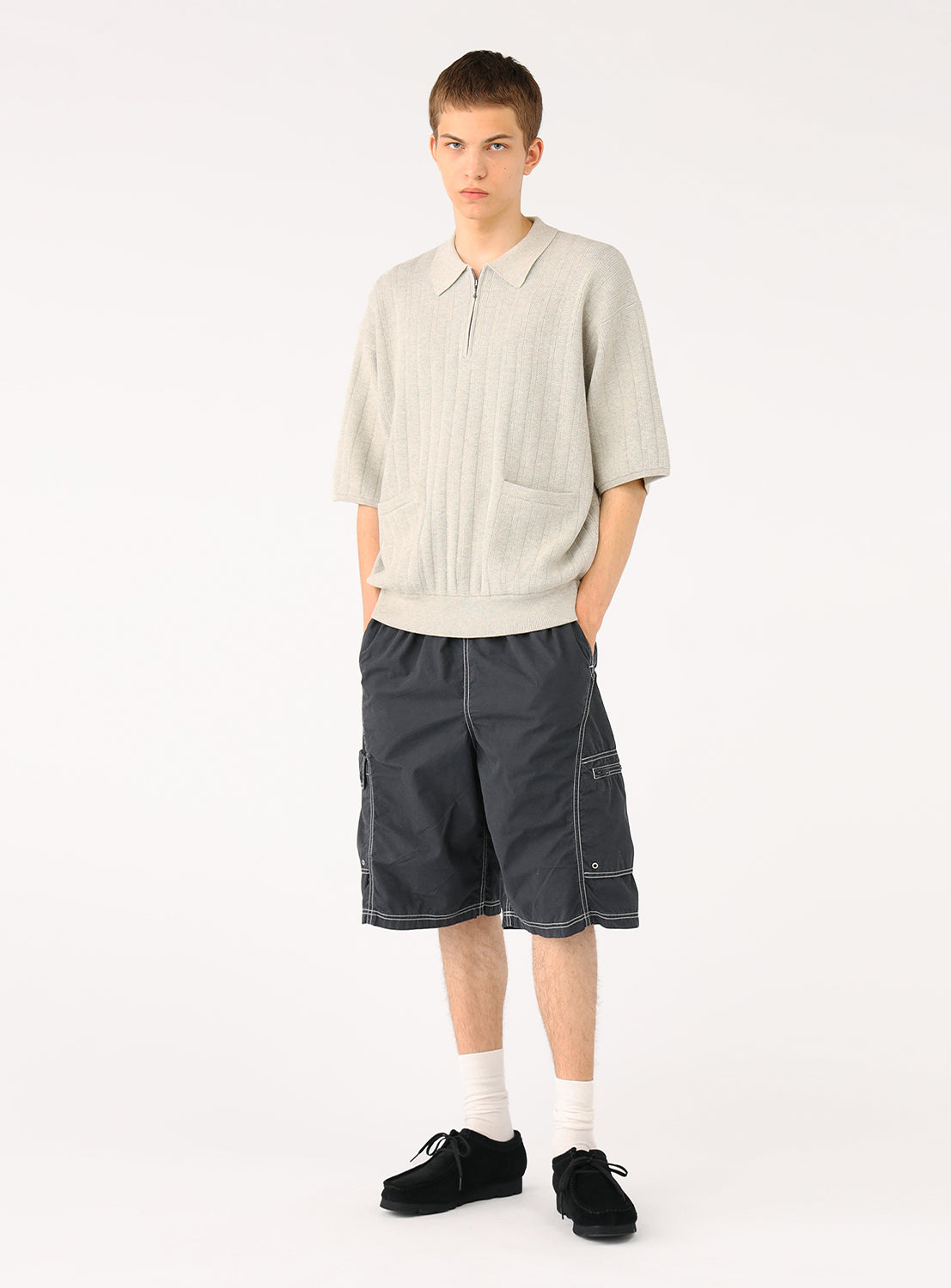 Male model in light grey knit polo and dark shorts, standing casually, full body view.