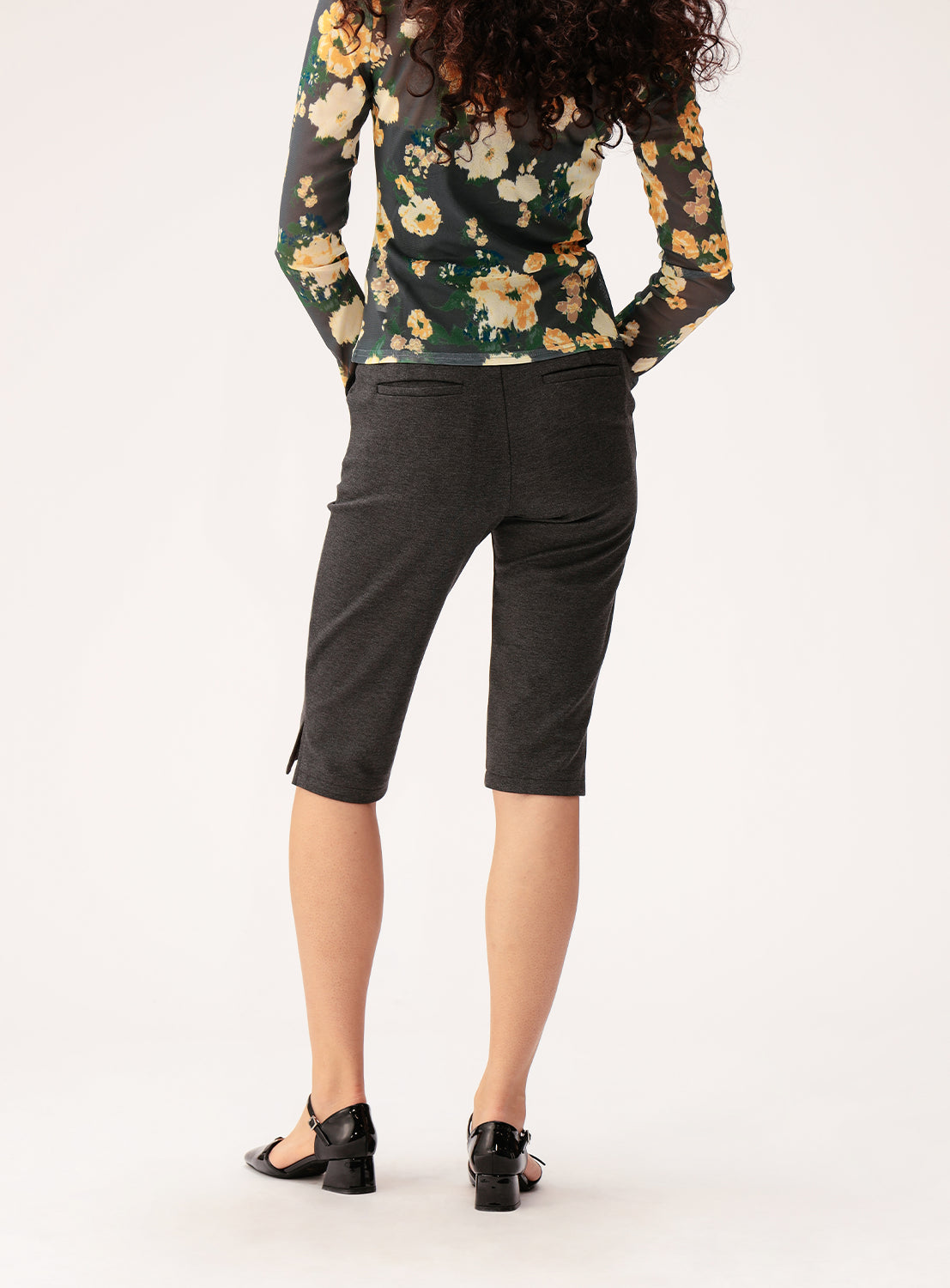 A person stands facing away, wearing a floral long-sleeve top, black Ray BEAMS Stretch Capri Pants with a slim silhouette, and black sandals. The background is plain and light-colored.