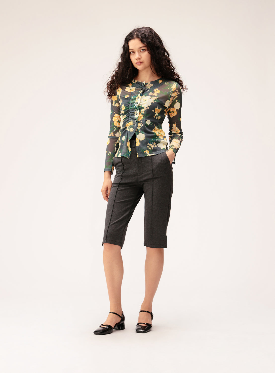 A woman with long curly hair wears a green floral top, Ray BEAMS Stretch Capri Pants in gray crafted from high-stretch fabric for a slim silhouette, and black Mary Jane shoes, standing against a plain light background.
