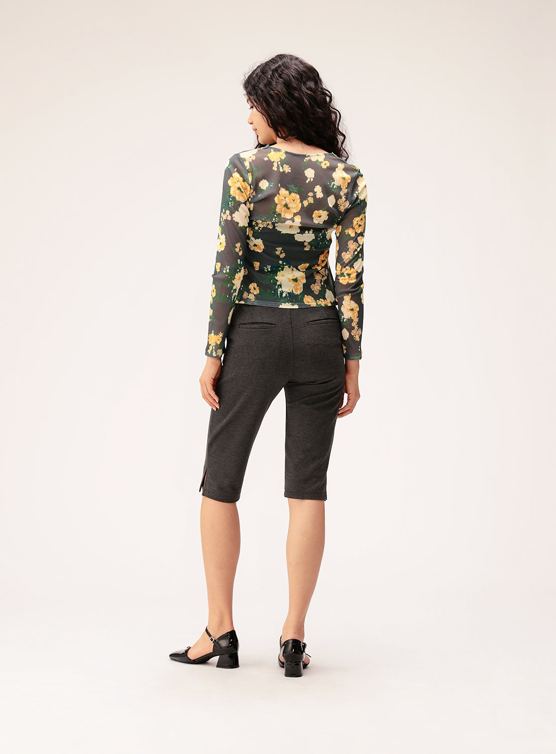 A woman with curly hair stands facing away in a neutral studio setting, wearing Ray BEAMS Stretch Capri Pants—dark high-stretch fabric capris—paired with black shoes and a sheer long-sleeve floral top.
