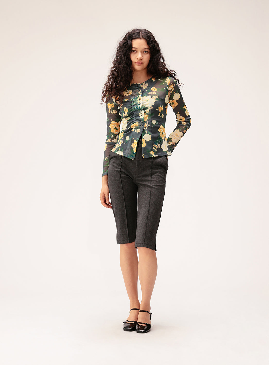 A woman with long curly hair wears a sheer floral long-sleeve top, Ray BEAMS Stretch Capri Pants in a slim silhouette crafted from high-stretch fabric, and black shoes, standing against a plain light background.
