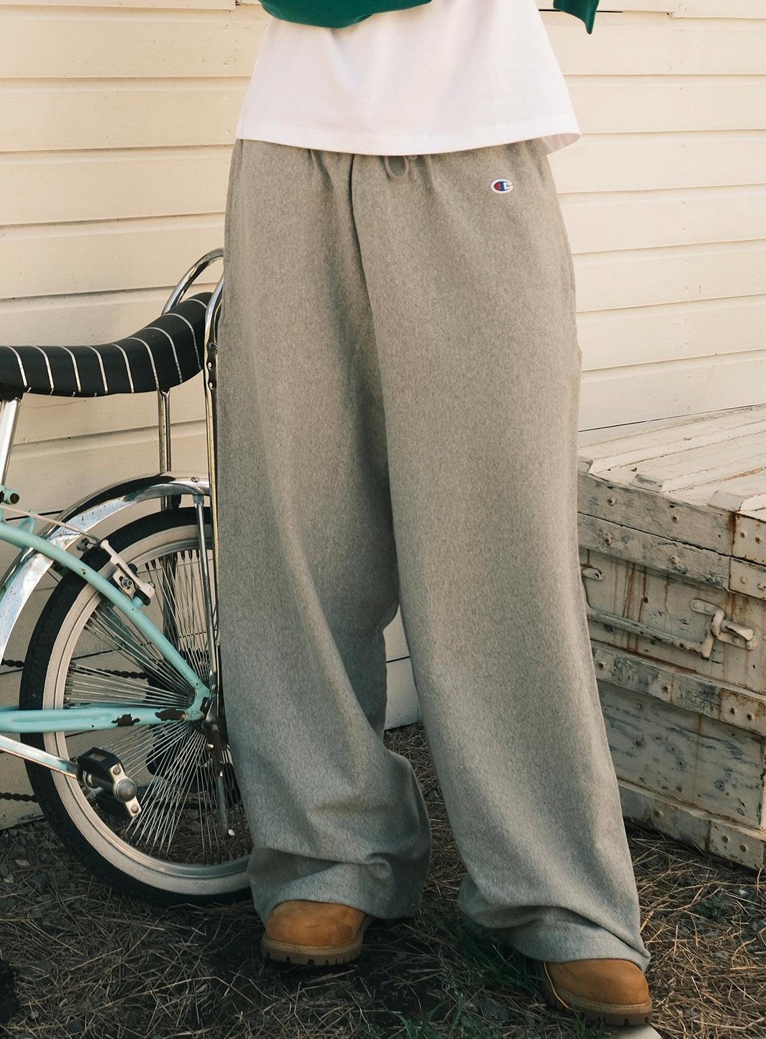 A person wearing loose gray [Exclusive] Champion Reverse Weave® Wide-Leg Pants from CHAMPION and tan boots stands next to a vintage bicycle and a wooden crate with a white wall in the background; only the lower half of their body is visible.