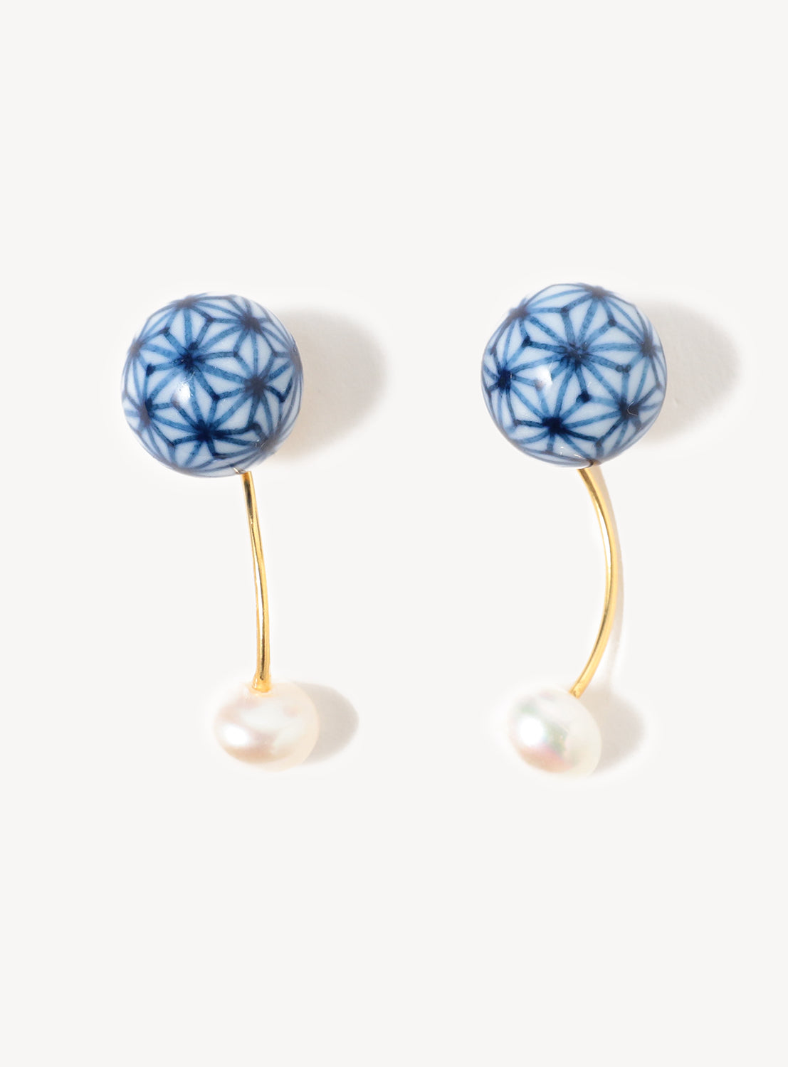The Hizen Jewelry Arita Ware Dangling Motif & Pearl Earrings feature blue and white patterned round Arita porcelain tops, elegant gold curved hooks, and delicate pearl-like drops, beautifully showcasing Japanese craftsmanship against a simple background.