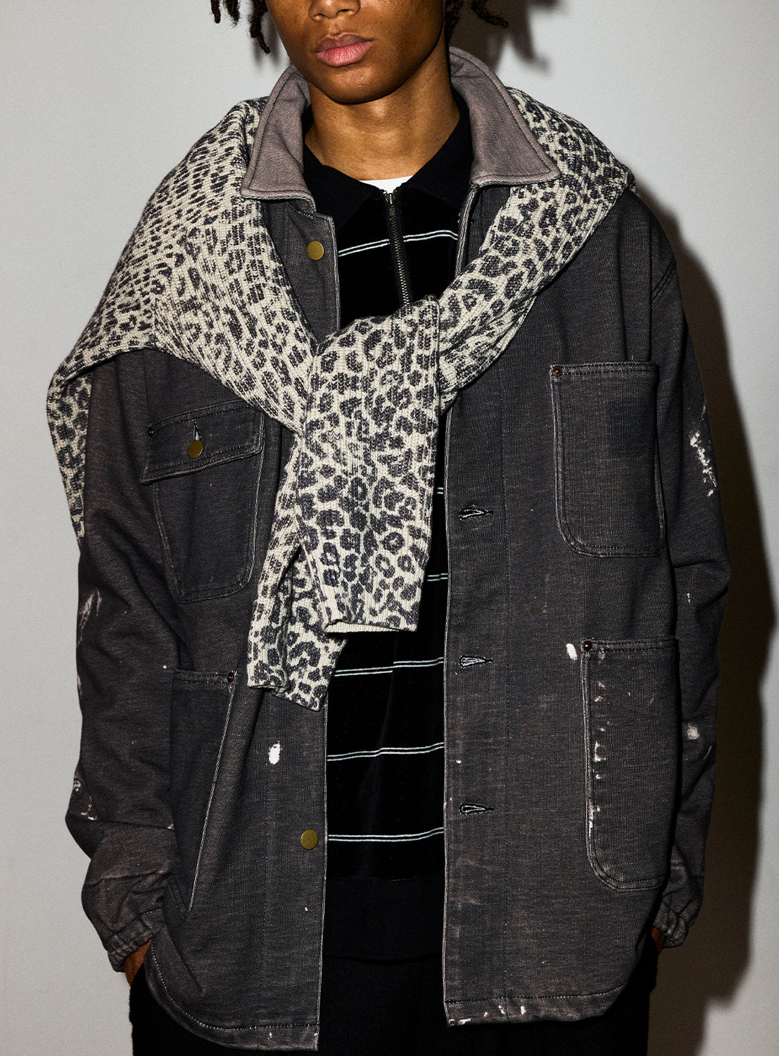 A person wears a relaxed fit, splash-bleached charcoal BEAMS Used Denim Coverall, a black and white striped polo shirt, and a leopard-print sweater draped over their shoulders.
