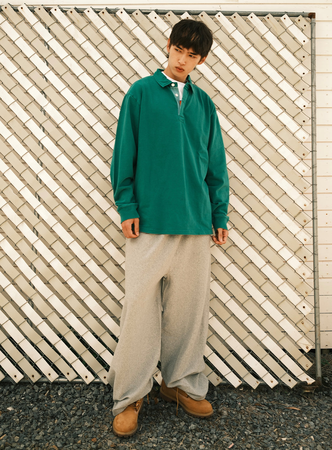 A person stands in front of a white lattice fence, wearing a green long-sleeve polo and CHAMPION [Exclusive] Champion Reverse Weave® Wide-Leg Pants, expertly crafted from sustainable American cotton, paired with tan boots and gazing slightly to the side on a gravel surface.