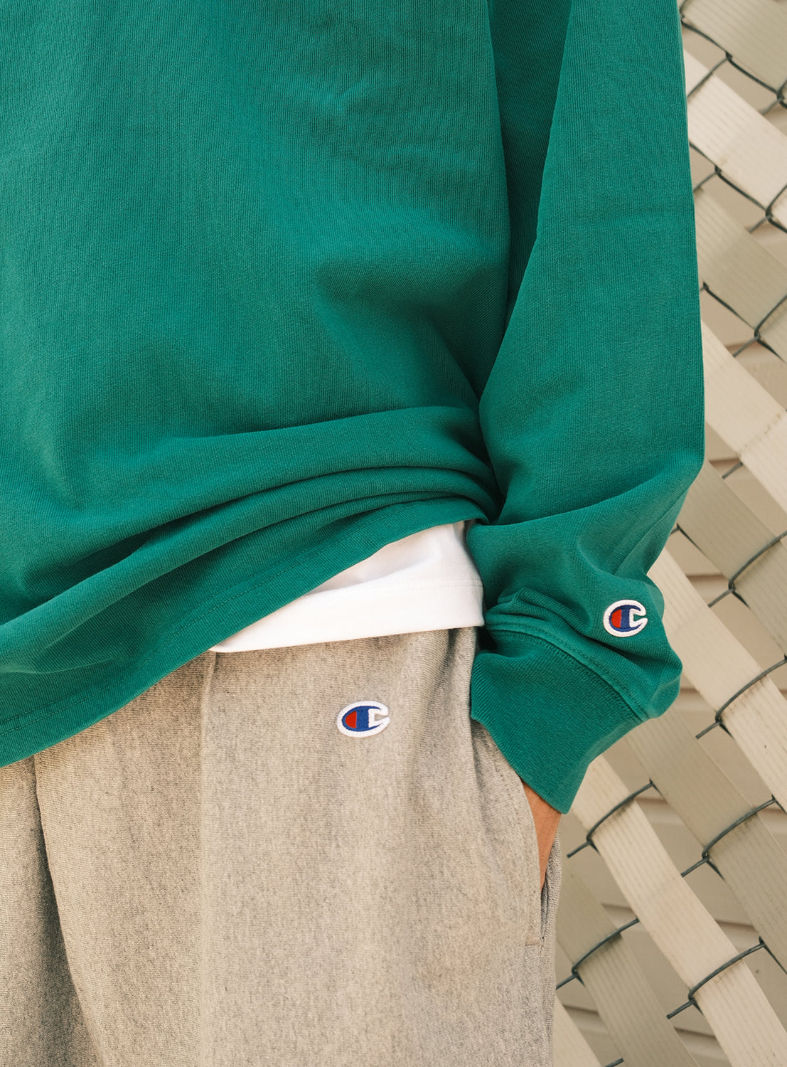 A person stands in front of a woven white fence, one hand casually in their pocket, wearing a green CHAMPION sweatshirt and gray sweatpants—effortlessly channeling the relaxed vibe of the [Exclusive] BEAMS x Champion Heavyweight Pigment-Dyed Rugby Shirt.