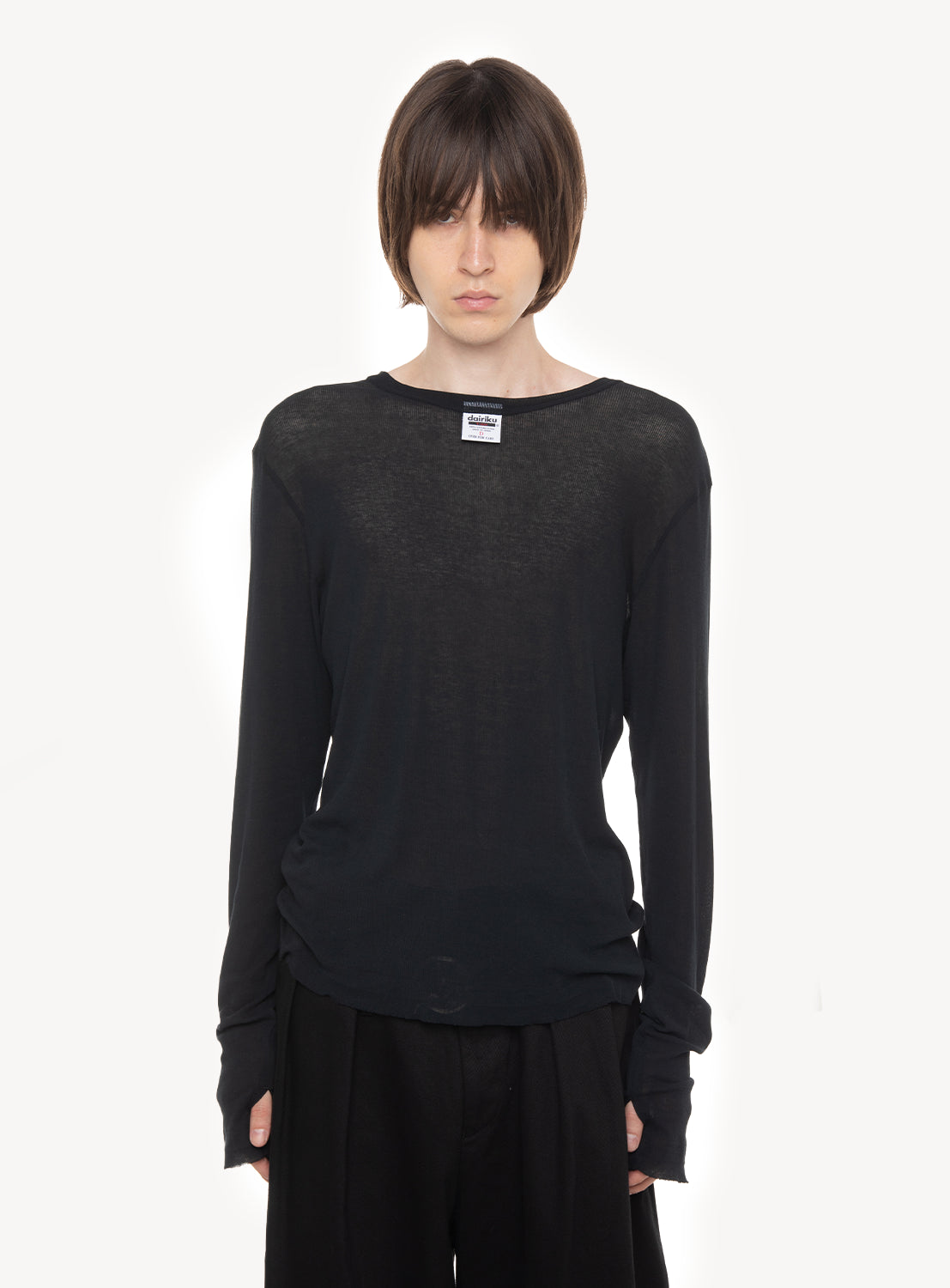 A person with straight brown hair stands against a plain white background, looking directly at the camera with a neutral expression. They are wearing the DAIRIKU “Daisy” Skelton Rib Tag Tee, layered underneath a sheer black long-sleeve shirt and paired with black pants.