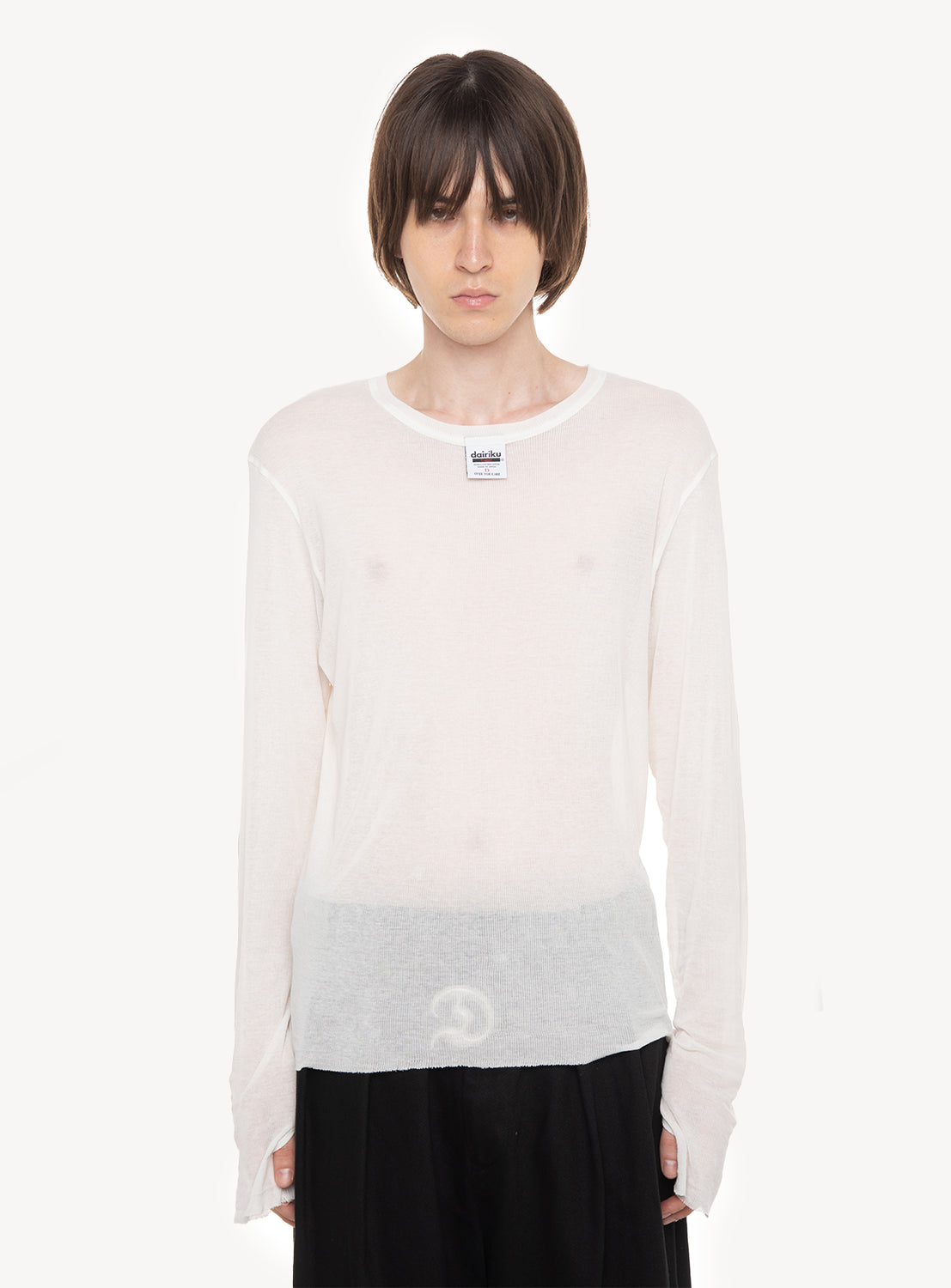 Wearing the DAIRIKU “Daisy” Skelton Rib Tag Tee, a person with short brown hair layers this sheer, long-sleeved white shirt over black pants, standing against a plain white background.