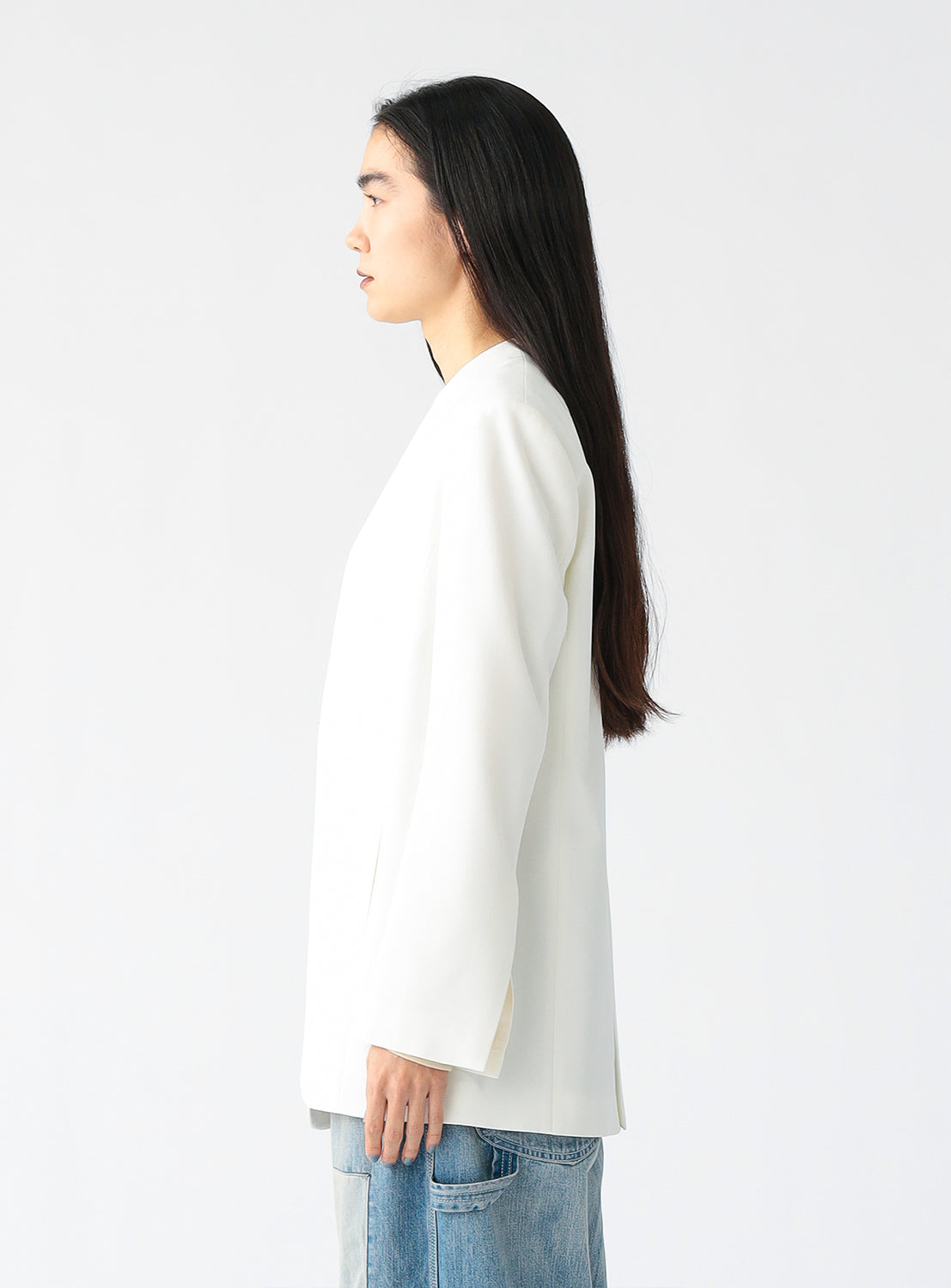 A person wearing a minimalist white blazer with a deep V-neckline and structured shoulders, styled over a sheer beige top and paired with light blue patchwork jeans, standing against a plain white background.