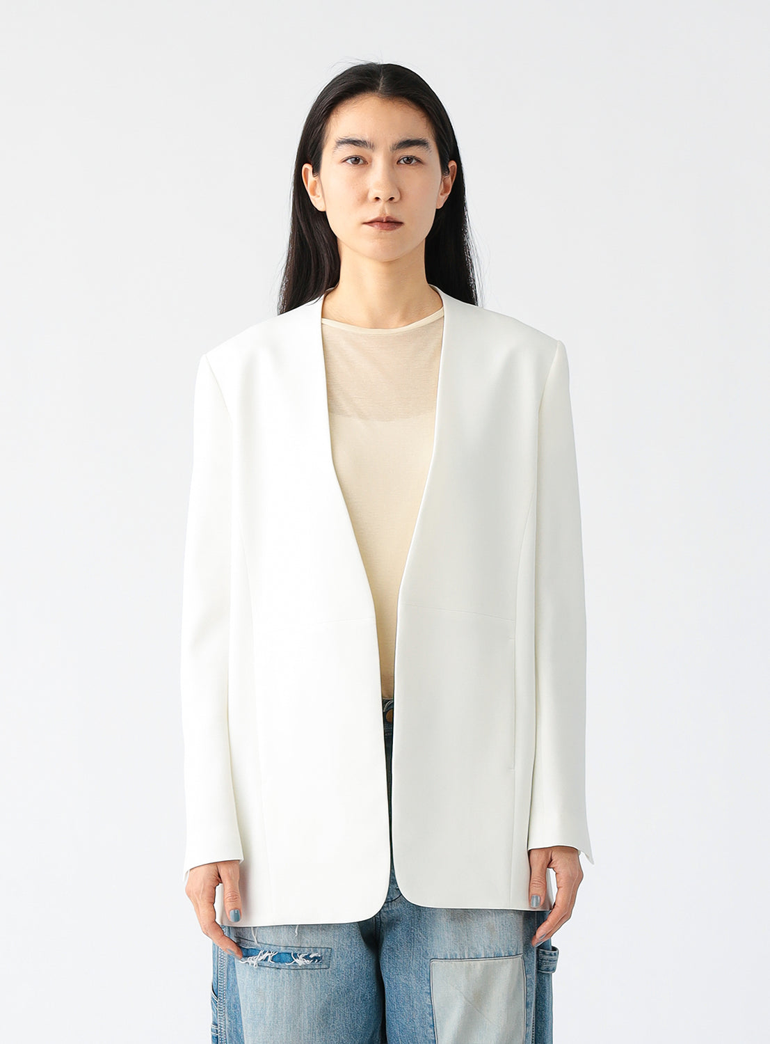 A person wearing a minimalist white blazer with a deep V-neckline and structured shoulders, styled over a sheer beige top and paired with light blue patchwork jeans, standing against a plain white background.