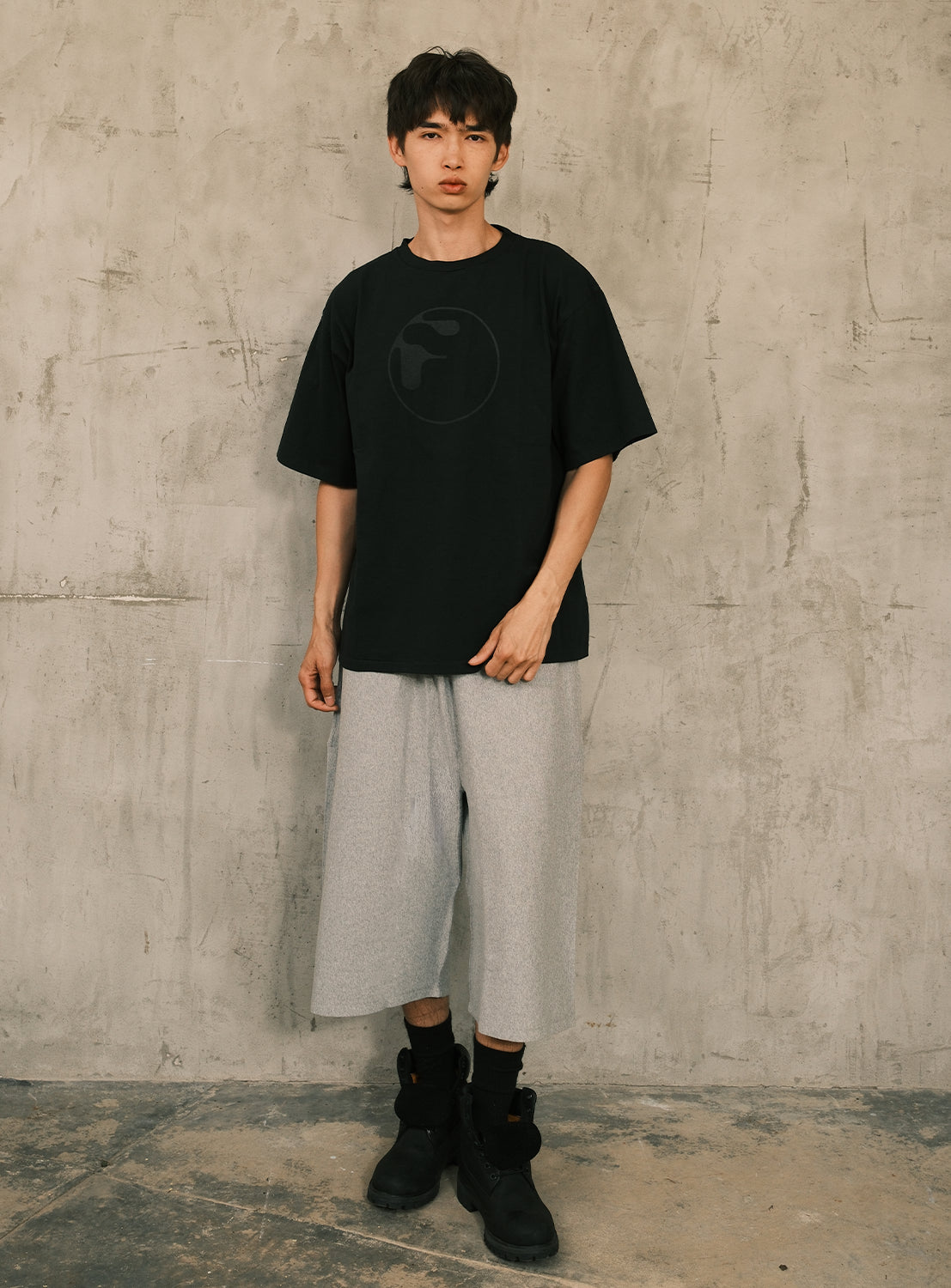 A young person stands against a textured concrete wall, wearing a loose black T-shirt with a subtle logo, [Exclusive] Champion Reverse Weave® Wide-Leg Shorts in gray by CHAMPION that fall to the knee, and black boots. The overall look is casual and modern.