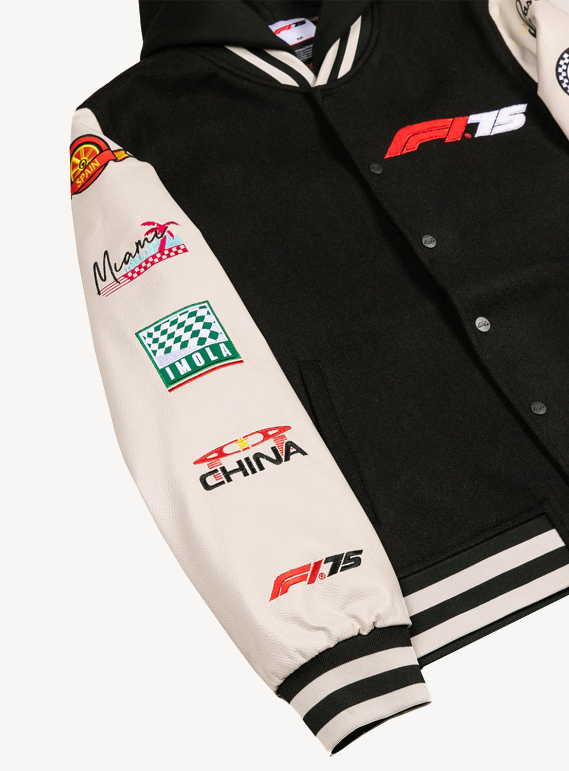 Close up of black button down varsity hooded jacket with white sleeves and racing locations going down the sleeves and the F1 75 logo on the chest. 