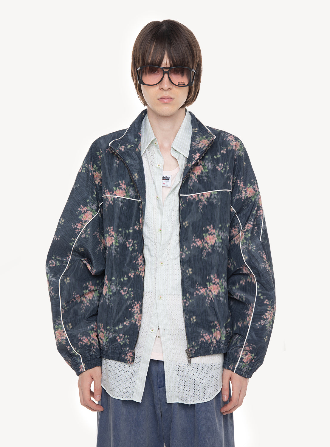 A person with short brown hair, wearing large sunglasses, the DAIRIKU “Fleur” Training Jacket over a light button-up shirt, and loose blue pants, standing against a plain white background.