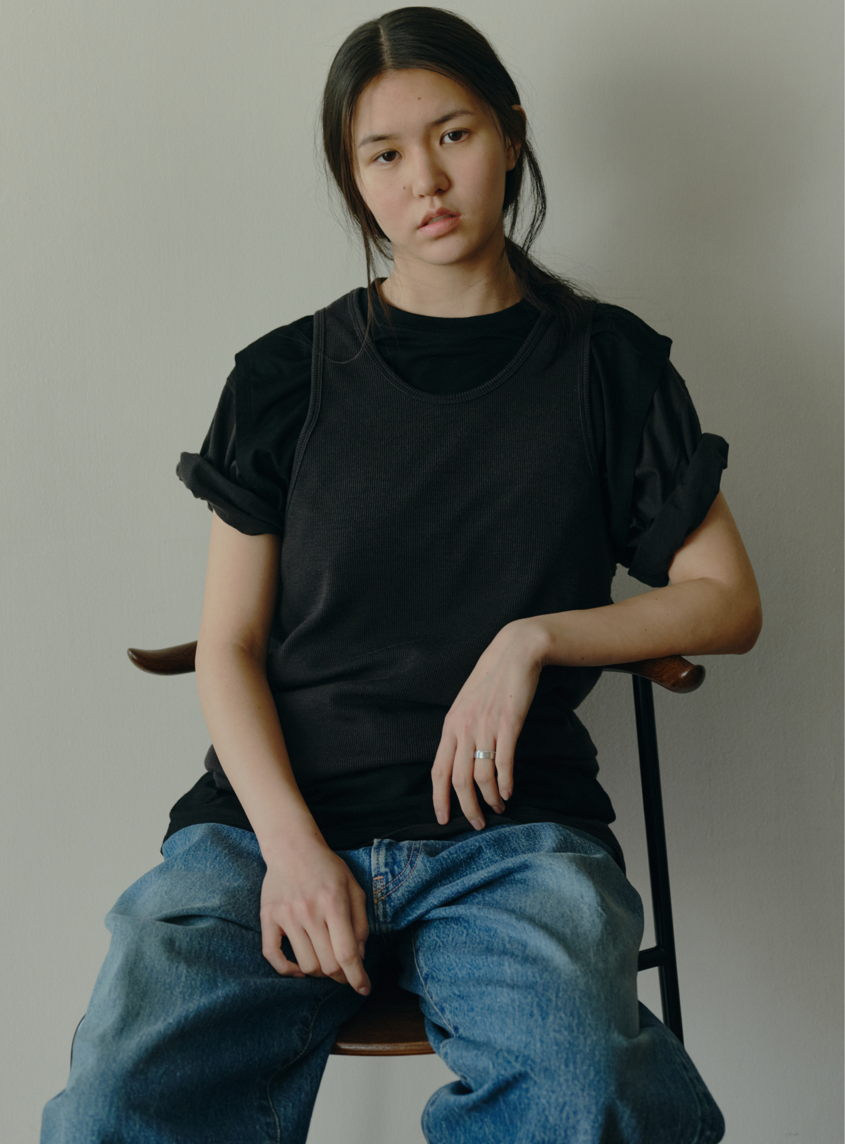 A person with long dark hair in a low ponytail sits on a chair against a plain wall, wearing BEAMS' the A - Washable Ribbed Wool Tank Top and loose blue jeans, looking at the camera with a neutral expression.