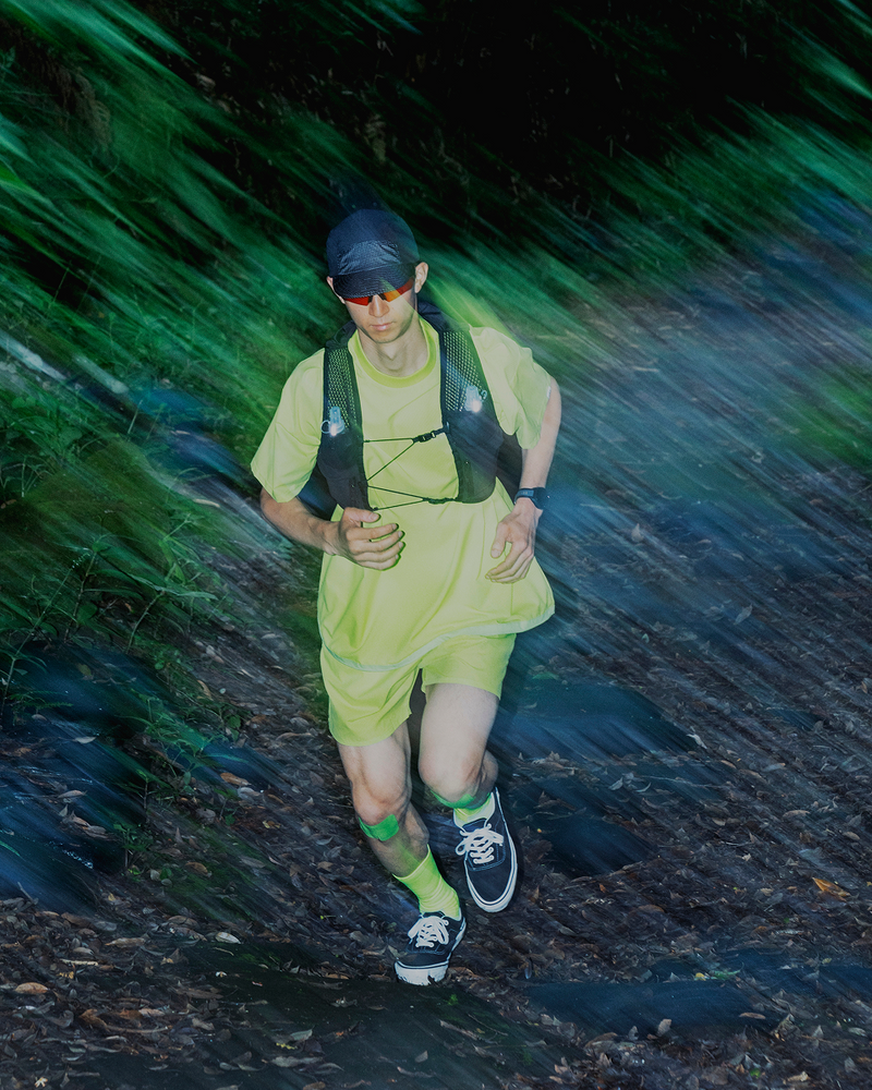 A person in neon yellow running gear and sunglasses runs through a forest trail, surrounded by motion blur and streaks of green and blue colors.