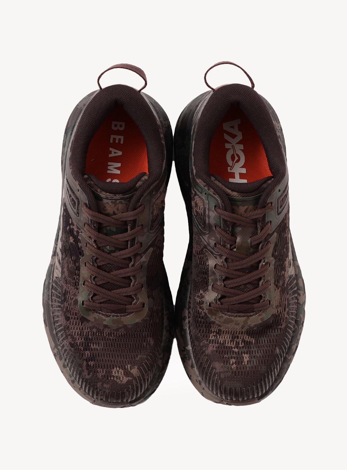 Top view of a pair of brown camo Hoka U BONDI 7 BEAMS sneakers with thick soles, brown laces, and orange insoles featuring “BEAMS” on the left and “HOKA” on the right. Designed with an engineered mesh upper for breathability and lightweight cushioning for all-day comfort, both shoes have fabric pull tabs at the heel.