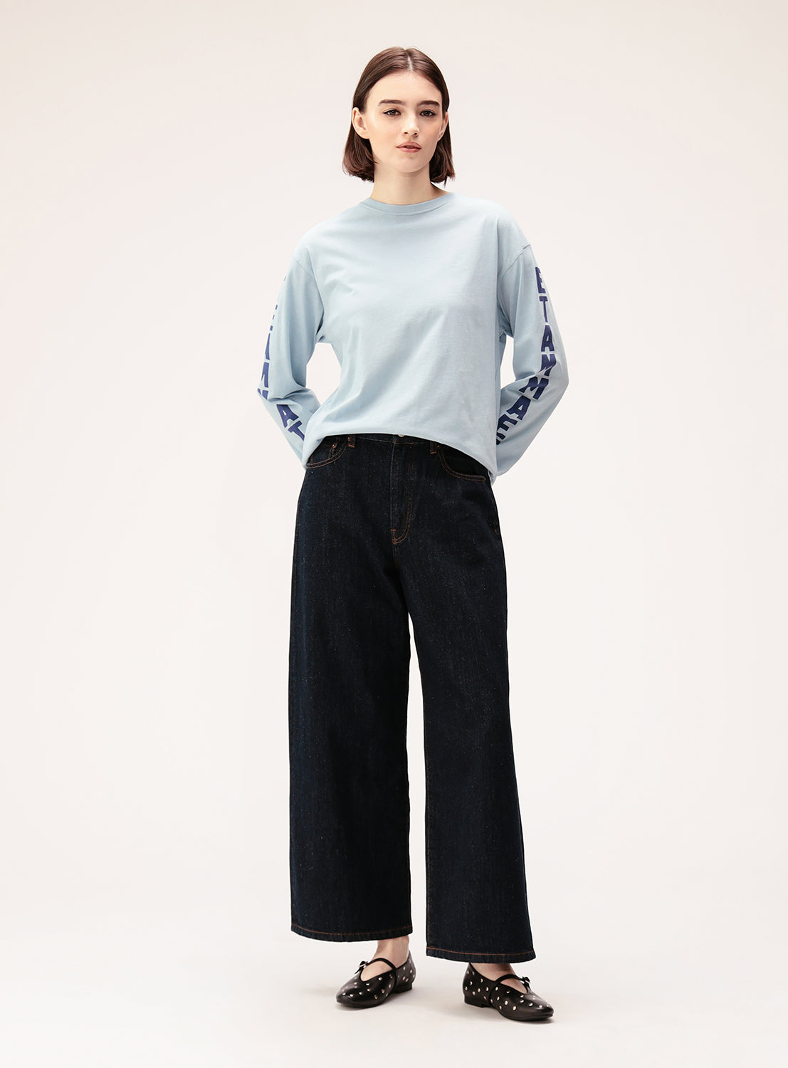 A woman stands facing forward, wearing the BEAMS BOY Big Logo Long-Sleeve T-Shirt in light blue with oversized sleeve logos, styled with wide-leg dark jeans and black cut-out detail shoes for a relaxed fit ensemble.