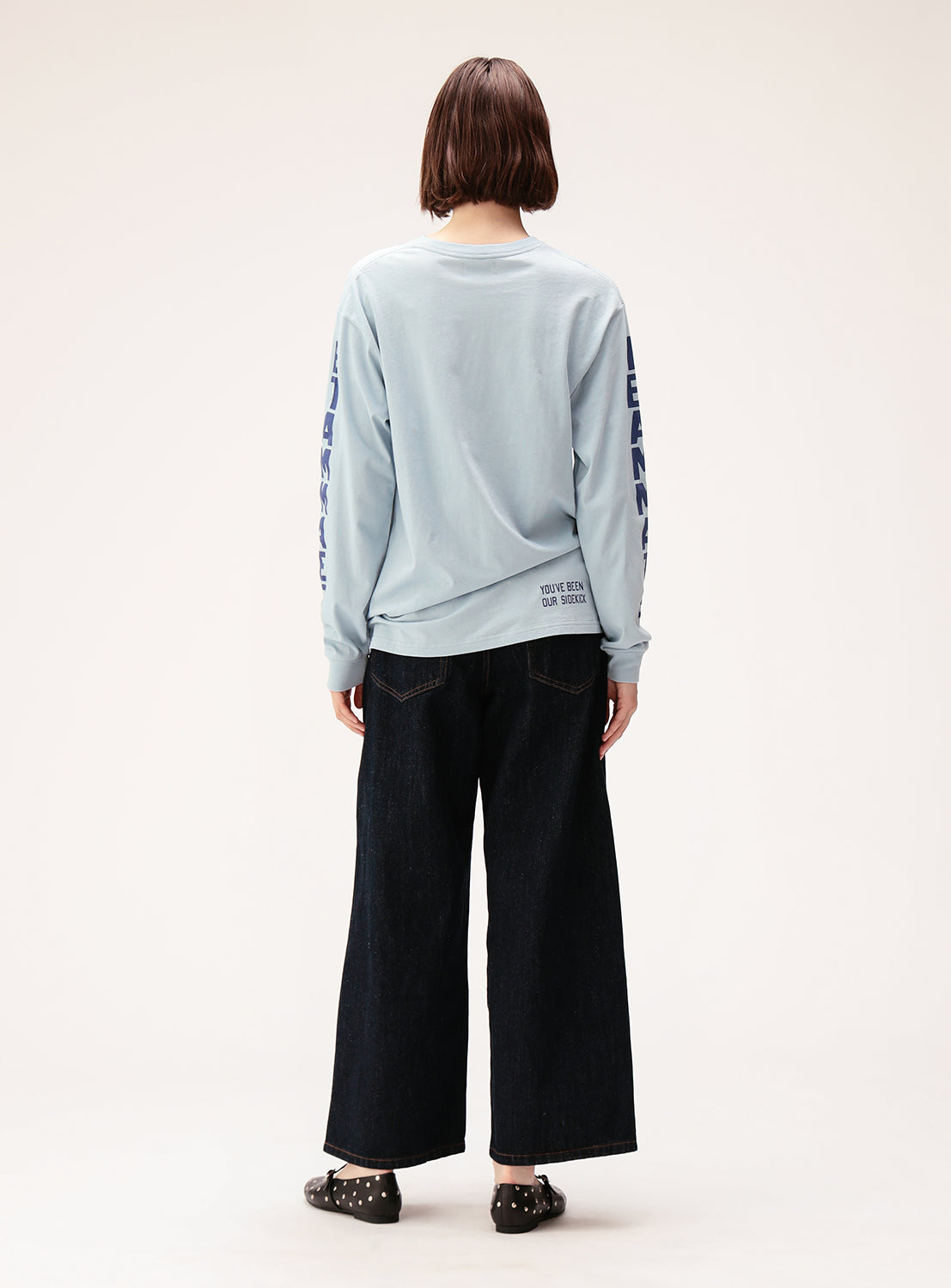 A person with short brown hair stands facing away against a plain background, wearing the BEAMS BOY Big Logo Long-Sleeve T-Shirt in a relaxed fit light blue style with oversized sleeve logos, paired with wide-leg dark jeans and studded black shoes.