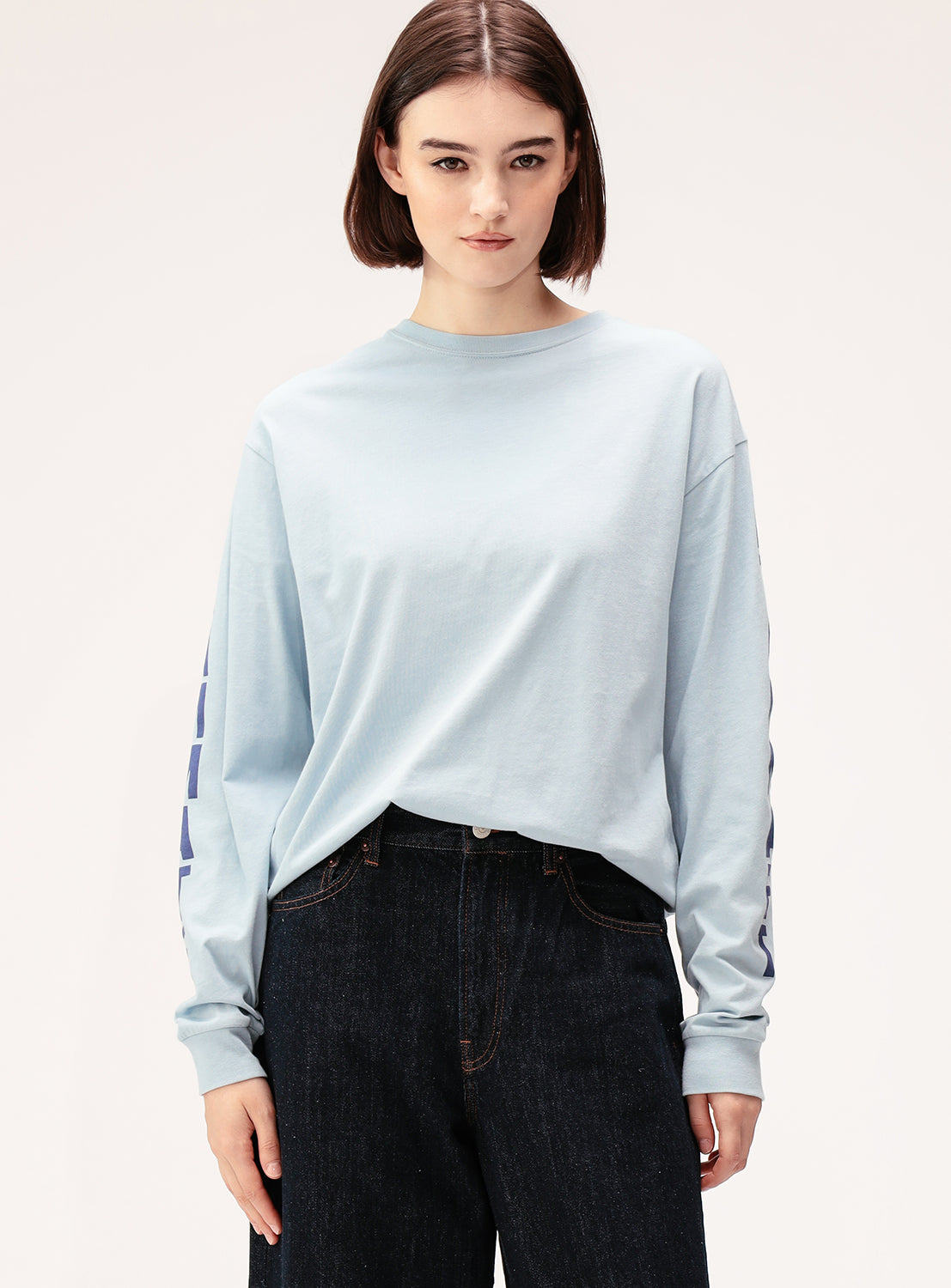 A woman with short brown hair wears the BEAMS BOY Big Logo Long-Sleeve T-Shirt in light blue, styled in a relaxed fit with dark pants. She faces forward with a neutral expression against a plain background.
