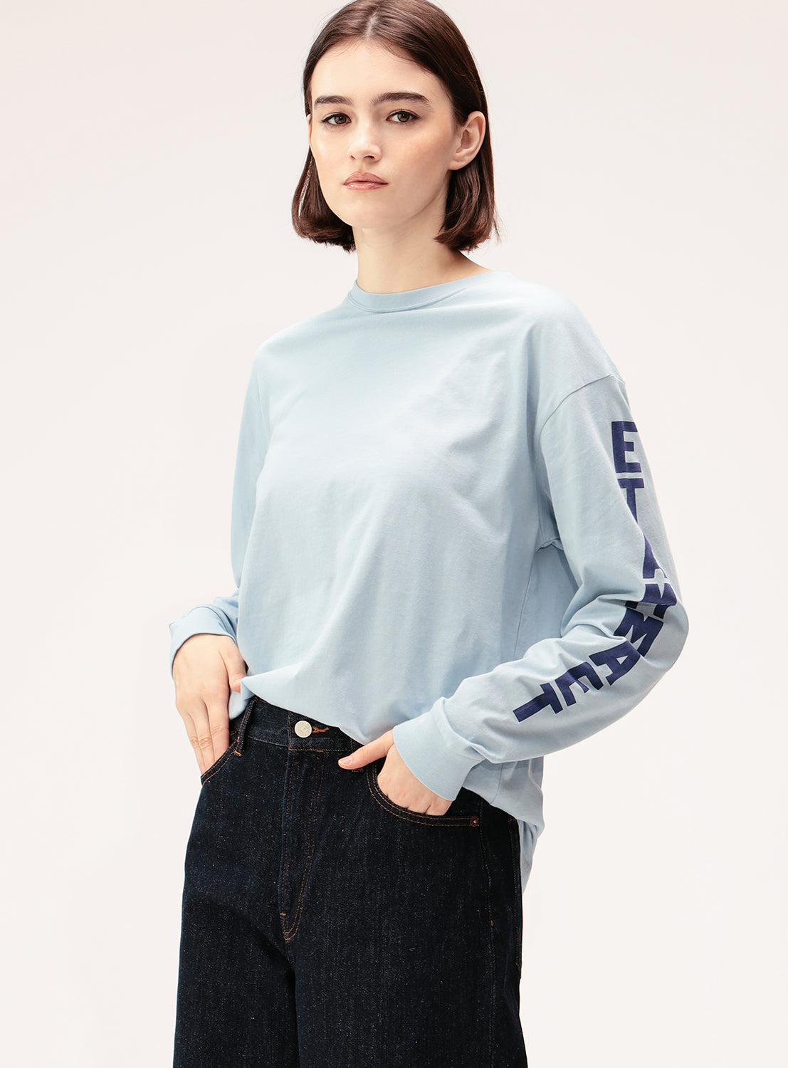 A young person with short brown hair wears the BEAMS BOY Big Logo Long-Sleeve T-Shirt in light blue, featuring oversized sleeve logos, paired with dark jeans and standing with a neutral expression against a plain background.